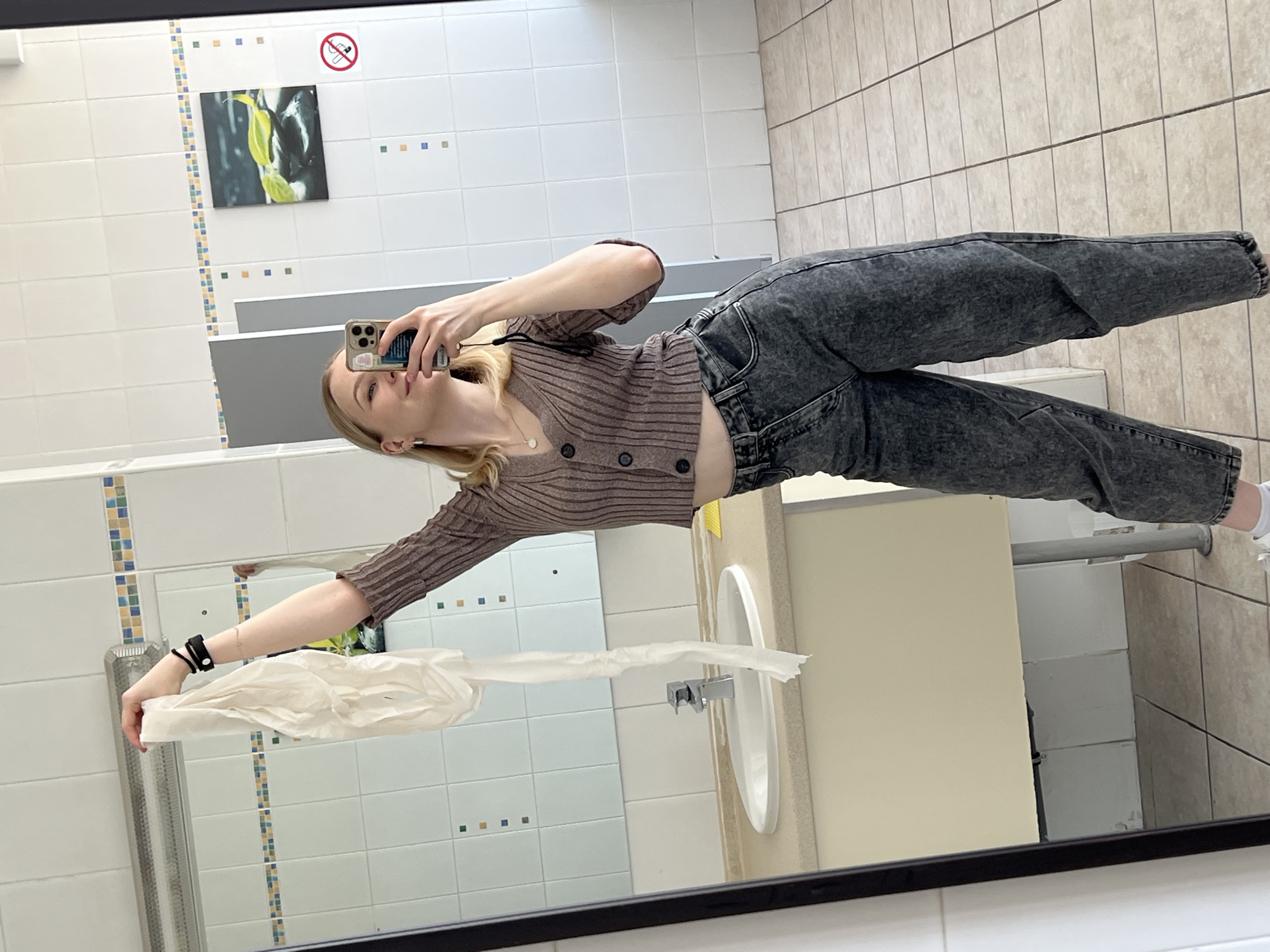 A young woman is taking a selfie in a public restroom. She has light skin and blonde hair and is wearing a brown, striped cardigan with buttons and dark-wash jeans. She is holding her phone up with her right hand and posing in front of a mirror. Her left hand is extended, holding a paper towel dispenser. The restroom has white tiled walls with decorative borders and a picture of a plant with a no-smoking sign above it. There is a white sink and countertop to the right of the mirror. The floor is tiled with beige squares. The photograph is taken from a slightly low angle, suggesting it's a mirror selfie. I am unable to verify the location in Khimki, Russia without additional context.