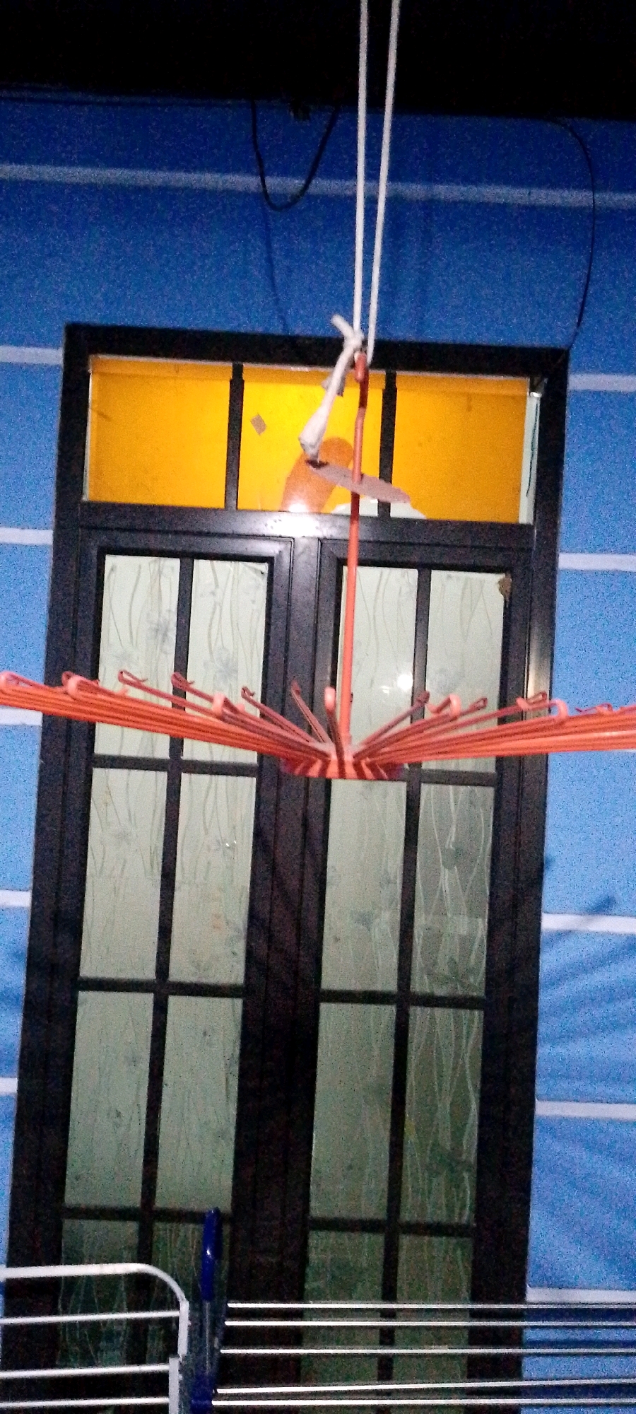 The image depicts an outdoor scene, likely in Sukamukti, Indonesia. A window with a dark frame and orange-tinted upper panes is visible. In front of the window, a pink drying rack with multiple rods is suspended by white ropes. The background is a blue textured wall with horizontal white stripes. In the lower portion of the image, part of a metal drying rack with horizontal bars is visible, along with a blue object that appears to be a clip. The scene is lit, suggesting it is daytime or artificial lighting is present.