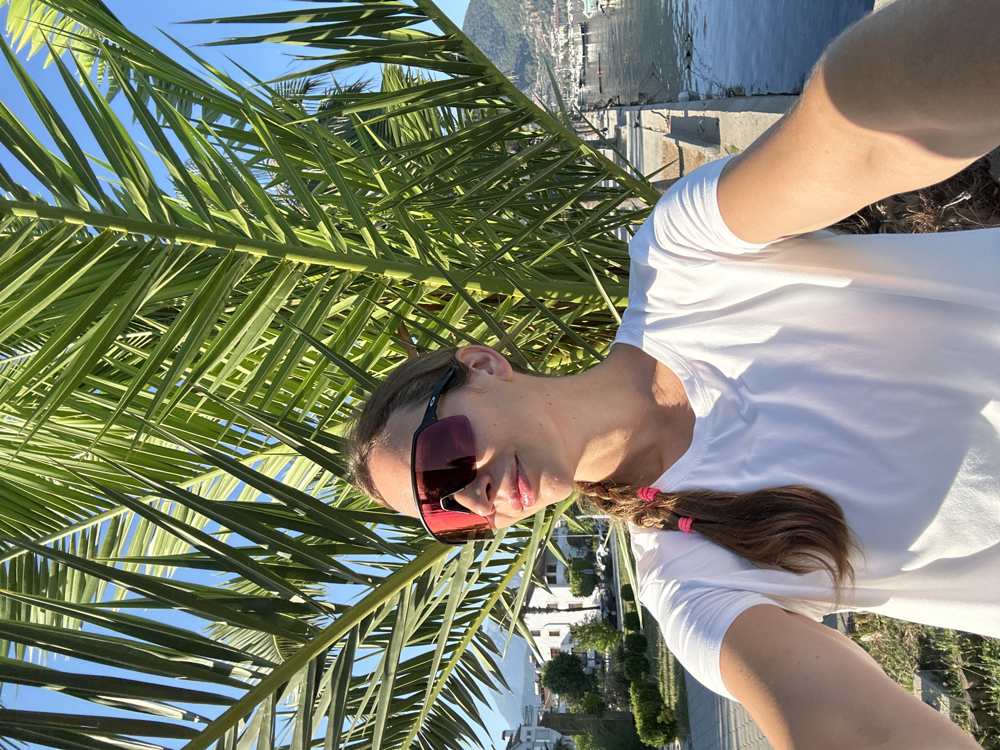 The image displays a female subject positioned in the foreground, capturing a selfie outdoors in Fethiye, Turkey. She has light skin, long brown hair styled in a side braid secured by two bright pink elastic bands. She wears large, dark-red mirrored wrap-around sunglasses with a black frame and a plain white short-sleeved t-shirt. Her head is turned slightly to the right, and her right arm is extended, holding the camera. Direct sunlight illuminates her face and arm.
A large palm tree with vibrant green fronds dominates the central and upper background, with clear blue sky visible between the fronds. To the left of the subject, behind the palm foliage, stand several multi-story white buildings featuring red-tiled roofs. A paved area or road is visible in front of these buildings, alongside green shrubbery. In the far distance, a mountain range is present.
To the right of the subject, a body of water, consistent with a bay or sea, is visible. A concrete pier or dock extends along the waterline, where multiple boats, primarily yachts and smaller vessels, are moored. Further in the distance, additional buildings stretch along the coastline and up a hillside, backed by more mountains. The scene is illuminated by bright, clear sunlight.