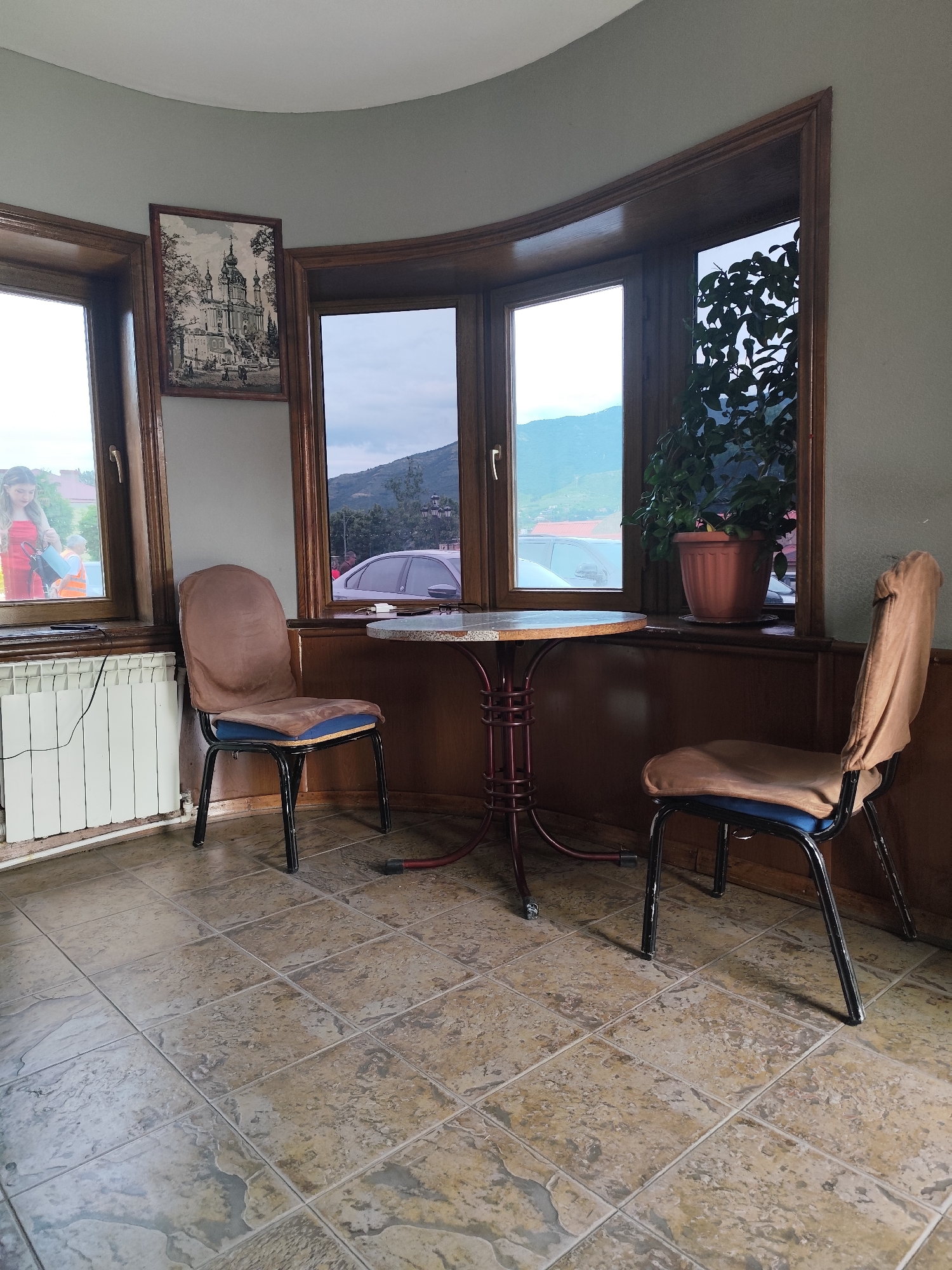 The image depicts an indoor seating area with a bay window offering a view of a hilly, overcast landscape. The room features a light grey wall and a wooden paneled lower section. In the foreground, a round, dark red metal-legged table with a light-colored top stands between two chairs. The chairs have black metal frames and light brown fabric upholstery with blue seat cushions. To the left, a white radiator is visible beneath a window where a young woman in a red dress is partially visible, looking down at something she is holding. To the right of the table, a large potted green plant is situated on the windowsill. The floor is tiled with large, irregular-shaped tiles in shades of beige and grey. Outside the bay window, cars and distant buildings are visible. A framed black and white print of a large architectural structure, possibly a church or cathedral, is mounted on the wall above the left window. The scene appears to be a cafe or restaurant in Mtskheta, Georgia, given the architectural style and the general landscape.