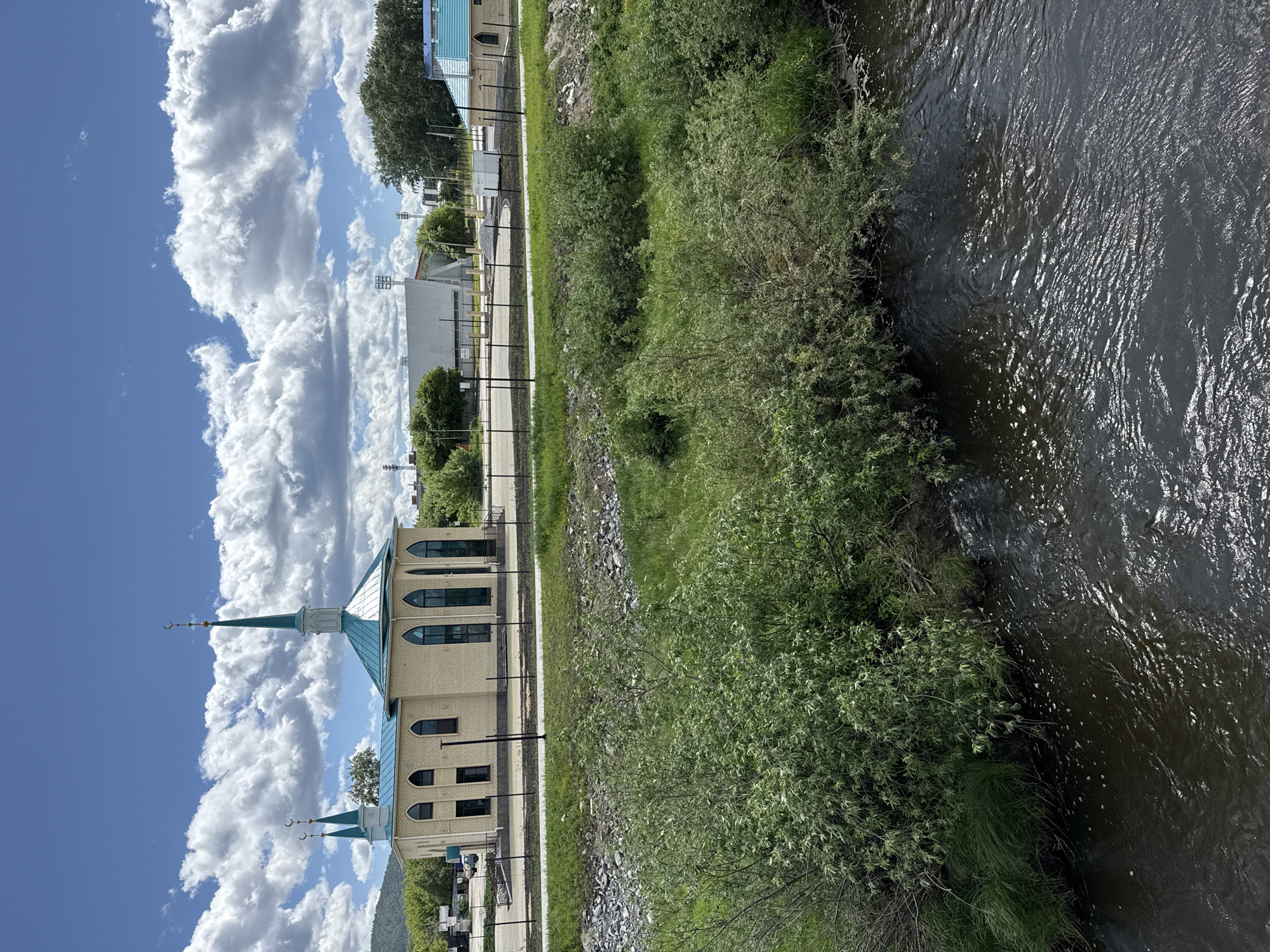 The image depicts a landscape featuring a river, a mosque, and adjacent infrastructure in the city of Miass, Russia.
On the left side of the frame is a tan brick mosque with a green roof and spires, set against a backdrop of a cloudy sky. A low fence runs along a pathway adjacent to the building.
To the right of the mosque, a grassy bank descends towards a river. The river takes up the right side of the frame, with its water appearing dark and reflecting sunlight. The riverbank is lined with green vegetation, bushes, and tall grasses.
In the background, partially obscured by the mosque and foliage, other buildings and structures are visible, including what appears to be a stadium and low-rise buildings.
