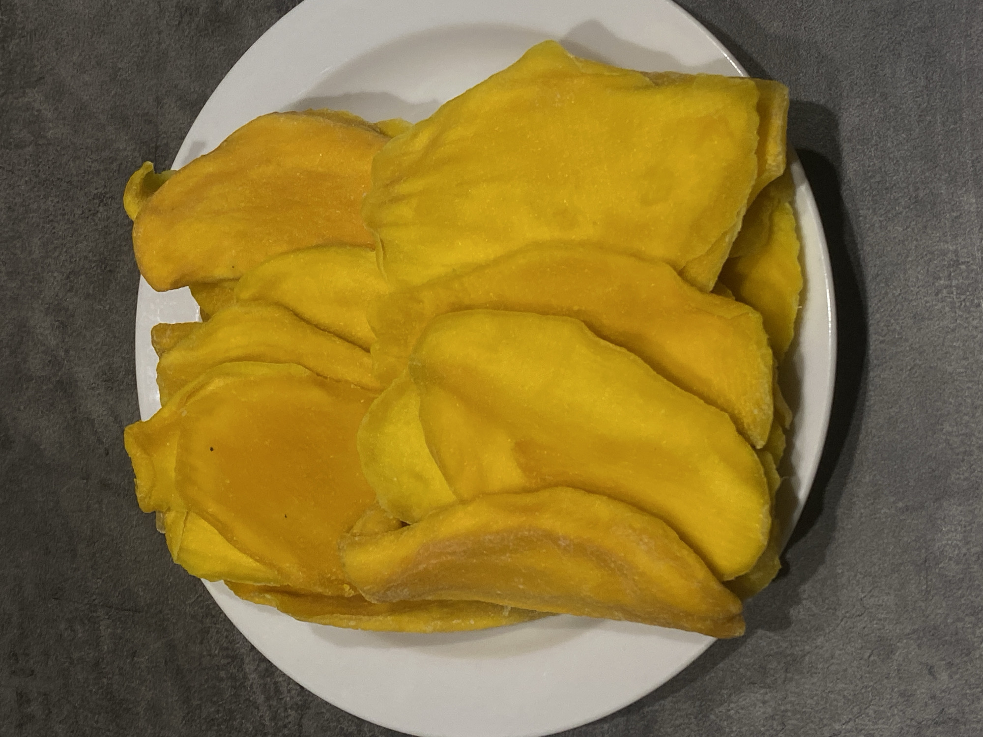 A white plate is filled with pieces of dried mango. The plate sits on a grey surface. The location is not discernible from the image.