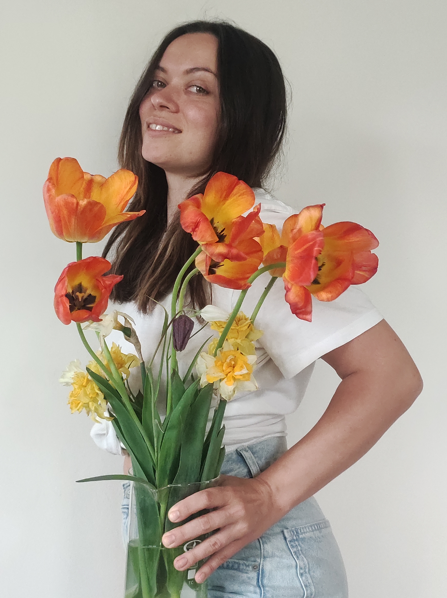 A young woman with long dark hair is holding a clear glass vase filled with orange and yellow flowers, including tulips and daffodils. She is wearing a white t-shirt and light blue jeans. The background is a plain white wall. The location is unknown.