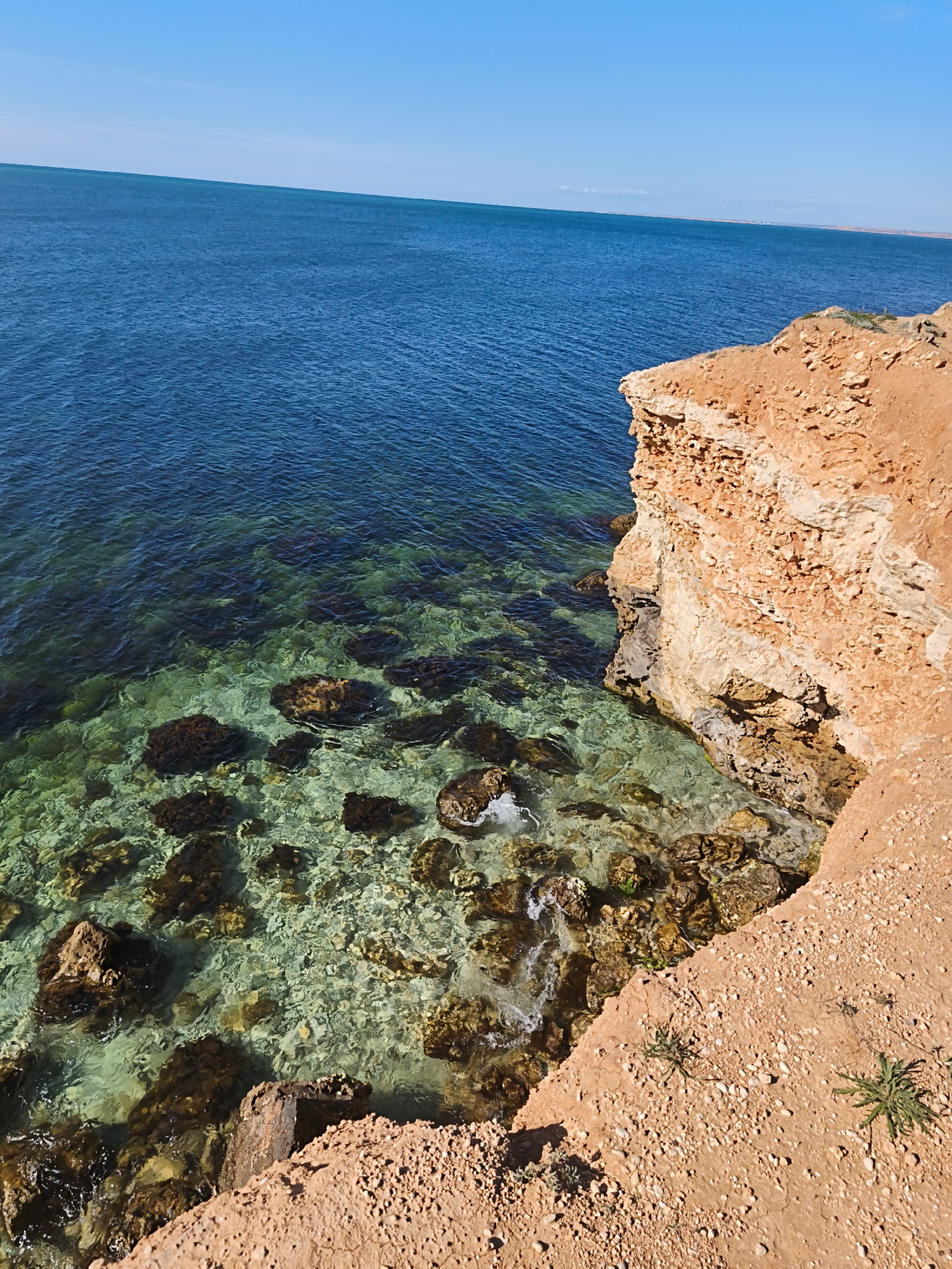 This media file captures a vivid coastal scene in Sevastopol, Ukraine, showcasing the clear waters of the Black Sea and rugged cliffs under a bright sky.
**Subjects:** The primary subjects are the expansive blue sea, a dramatic reddish-brown cliff face, and the clear, rocky seabed near the shore. The sea exhibits a beautiful gradient, transitioning from a translucent turquoise and light green near the coast, where submerged rocks are clearly visible, to a deep, rich blue further out. The cliff on the right side of the image is made of eroded, layered rock, showing textures and crevices. Scattered small green plants can be seen growing on the sandy-rocky ground at the top and edge of the cliff. The sky above is a clear, uninterrupted expanse of bright blue.
**Activity or event taking place:** No specific activity or event is depicted. The scene is a static landscape view of a natural coastal environment. Small waves can be seen gently lapping against the rocks and base of the cliff.
**Notable details:** The time of day appears to be bright daylight, possibly late morning or early afternoon, indicated by the vibrant colors and strong, direct sunlight creating clear visibility and defining textures. The weather is perfectly clear and sunny, with no clouds in the sky. Location cues include the distinctive color of the water, the rocky coastline, and the specific type of eroded cliff, consistent with coastal areas. The overall impression is one of natural beauty and tranquility. There is no visible text in the image.
