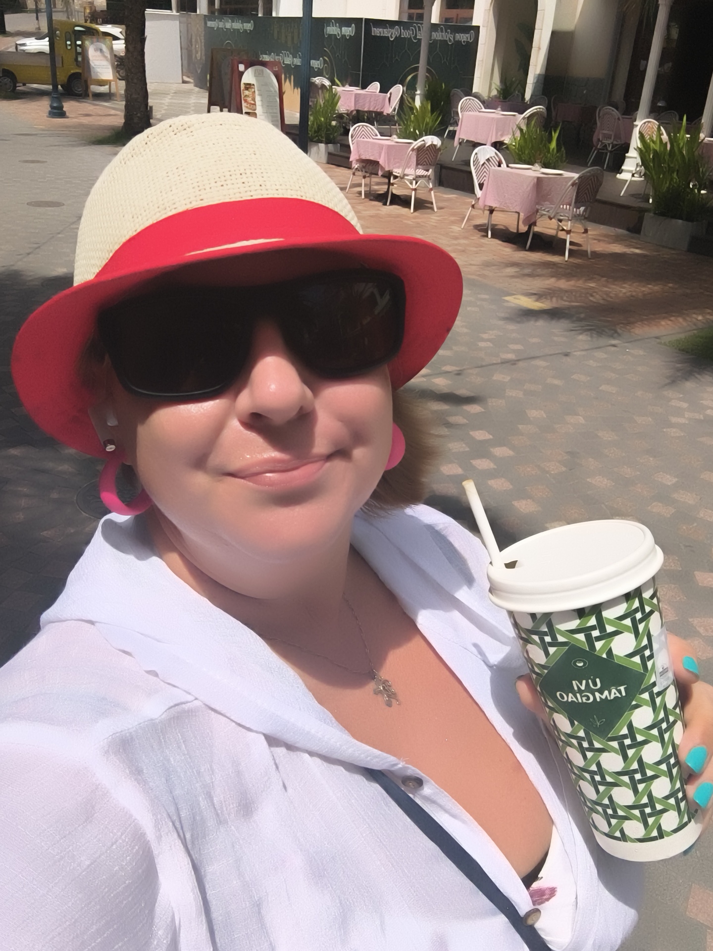 A woman in a red and white sunhat and sunglasses smiles at the camera, holding a paper coffee cup with a green geometric pattern and white lid. She is wearing a white open-collared shirt and has bright turquoise fingernails. A small silver necklace with a cross pendant is visible. She has large pink hoop earrings. The background shows an outdoor seating area of a cafe with tables and chairs, some covered with pink tablecloths. In the distance, a yellow vehicle and a palm tree are visible. The paving stones on the ground are light grey with darker grey diamond-shaped patterns. The scene appears to be in the city of Phú Quốc, Vietnam, suggesting a tropical, possibly coastal, environment.