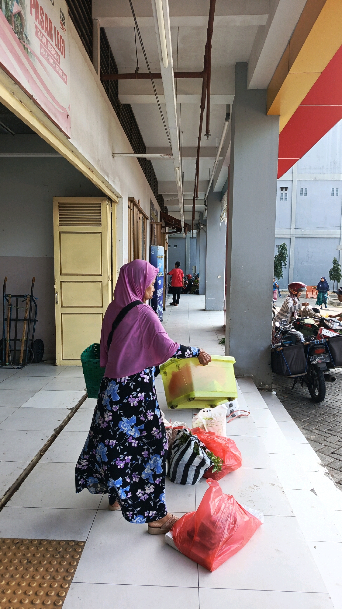 The image depicts an outdoor scene in Nologaten, Indonesia. In the foreground, a woman wearing a pink hijab and a floral dress is standing, holding a light green plastic container with wheels. In front of her and to her right are several shopping bags: one black and white striped, two red plastic bags, and a white bag with text. To her left is a green mesh basket. Behind her, there's a yellow door with horizontal slats and a metal cart with two wheels. Further in the background, a person in a red shirt is walking away, and other individuals are visible near a motorcycle with a sidecar parked on a paved area. The architecture includes a covered walkway with gray pillars and a white ceiling with fluorescent lights and exposed pipes. The facade above the walkway features sections of yellow and red. The ground is tiled with a white grid pattern, and a textured brown tactile paving strip is visible in the bottom left corner.