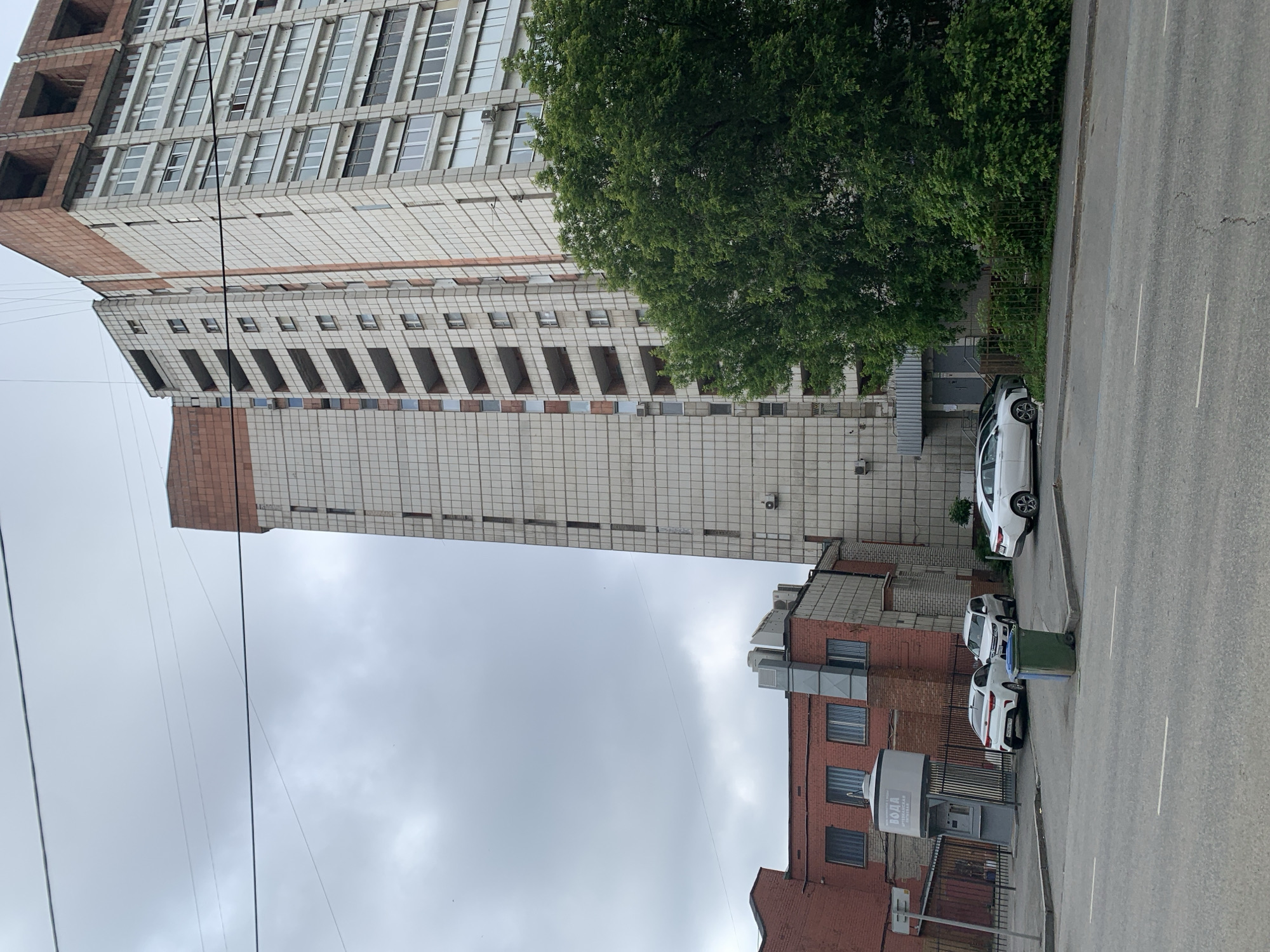 The image depicts a street scene in Perm, Russia, under an overcast sky. Dominating the right side of the frame is a tall, multi-story residential building with a pale gray facade, featuring numerous balconies and windows. The upper portion of this building appears to be under construction, with exposed brickwork and unfinished elements. To the left of the tall building is a lower, red brick industrial-style building with a metal roof and several windows. Adjacent to this is another section of the tall building, also clad in pale gray tiles. A lush green tree with dense foliage is situated in front of the tall building, partially obscuring the lower floors and an entrance. In the foreground, a paved road runs horizontally across the bottom of the image, with white lane markings. A sidewalk runs along the side of the road, where several white cars are parked, including a Kia sedan and a white SUV. A green trash bin is also visible on the sidewalk. A prominent feature on the sidewalk near the red brick building is a grey vending machine labeled "ВОДА" (Water). A black metal fence surrounds the area in front of the red brick building and the vending machine. Overhead power lines crisscross the sky. The overall impression is an urban environment with a mix of residential and industrial architecture on a cloudy day.