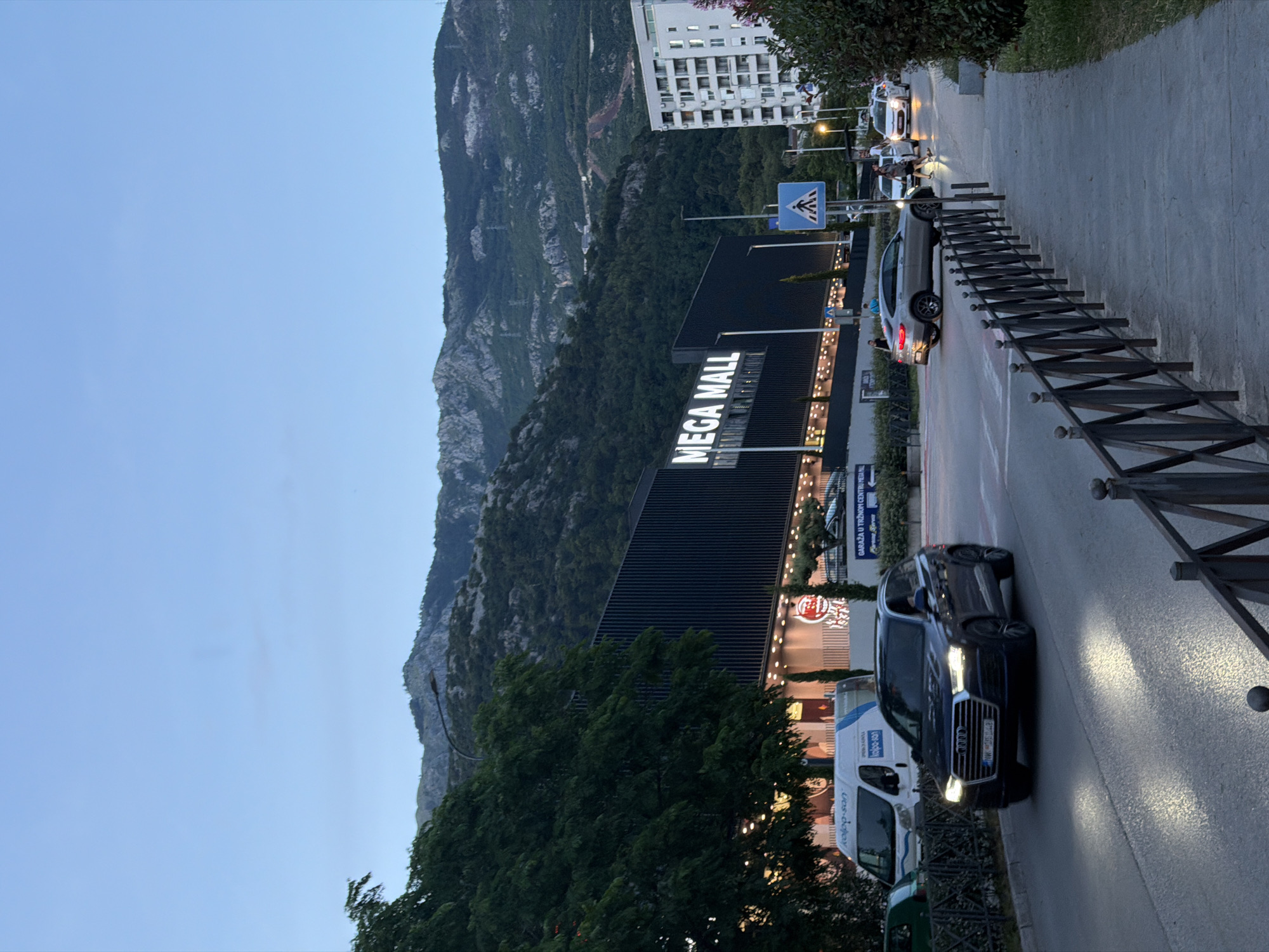 The image depicts an urban street scene in Budva, Montenegro, during dusk or early evening. A street, with several vehicles, runs horizontally across the frame. In the foreground, a dark Audi SUV with illuminated headlights is prominent, driving or parked on the right side of the road. Further down, a silver sedan and other cars are visible, and a white van with "kolpo-son" written on its side is parked.
A large, dark building with "MEGA MALL" prominently displayed in bright, illuminated white letters on its facade dominates the left side of the road. Above the mall, and stretching across the background, are significant, tree-covered mountains. To the right of the mountains, a multi-story white apartment building is visible.
A sidewalk runs along the right edge of the road, separated by a black metal fence. A blue pedestrian crossing sign is visible near the street. Pedestrians can be seen on the sidewalk. Other visible text includes "GARAŽA U TRŽNOM CENTRU MESA ML" on a sign near the mall. The sky is a clear, light blue, indicating the transition from day to night, with streetlights and building lights beginning to illuminate the scene.