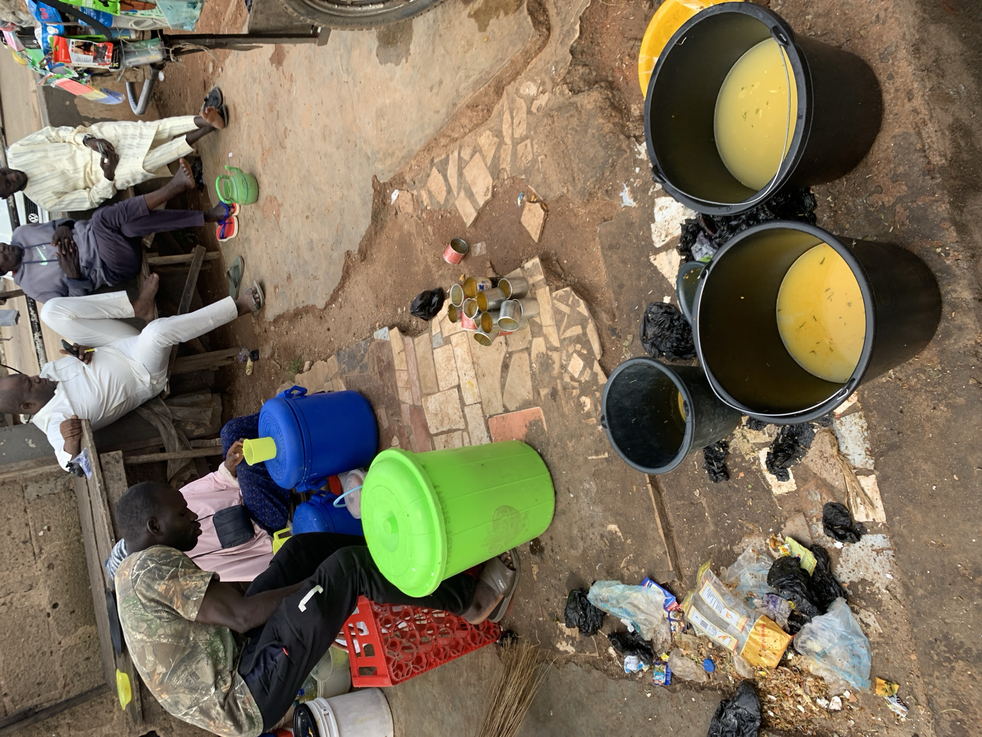 This image depicts a street scene in Jos, Nigeria, likely a market area, characterized by a dusty, uneven ground with scattered debris. In the foreground, two black buckets filled with a yellow liquid are prominent. To the left of these buckets, a man wearing a camouflage t-shirt and black pants is partially visible, bending down. Behind him, a large green plastic bin with a lid stands next to a red plastic crate. Further back, several people are seated on a wooden bench under a makeshift wooden structure, which appears to be a stall or vendor's setup. One man in a white traditional outfit is seated furthest to the right, holding a mobile phone. Another man in a grey hooded sweatshirt and dark pants is seated to his left, with his hands clasped. A person in a light pink garment is also seated behind the man in the camouflage shirt, with a black pouch on their lap. Scattered on the ground are various items, including cans, plastic bags, and what appears to be food waste, indicating a lively but potentially unkempt environment. The overall atmosphere suggests a bustling public space where daily commerce and social interaction take place amidst the city's urban landscape.