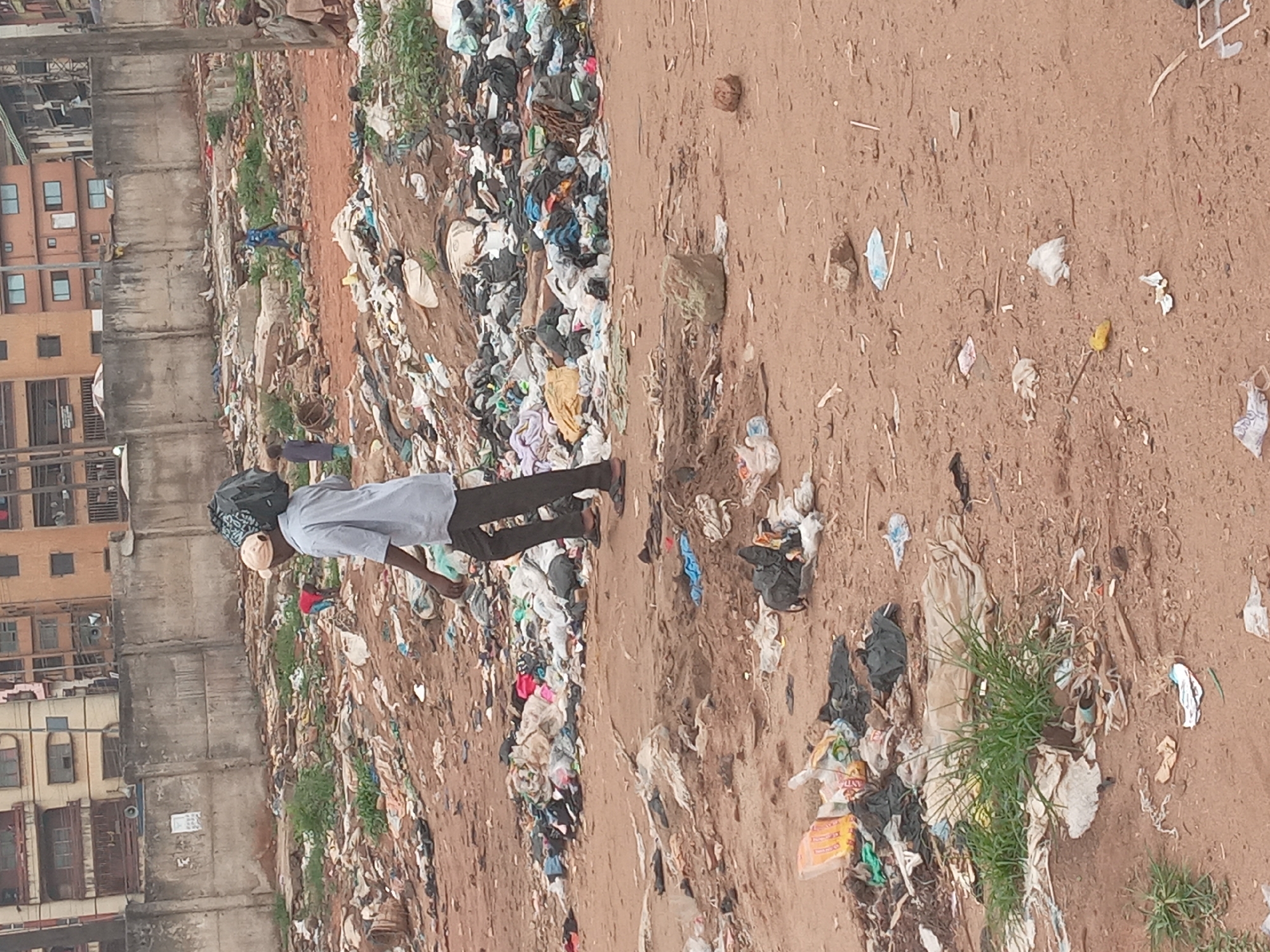 A man with a bag on his head walks on a dirt path between a pile of refuse and a concrete wall. The wall borders a multi-story residential building. The location is likely a slum area in Onitsha, Nigeria. The refuse is mostly plastic, fabric, and paper. More refuse litters the dirt path, intermixed with a few tufts of grass.