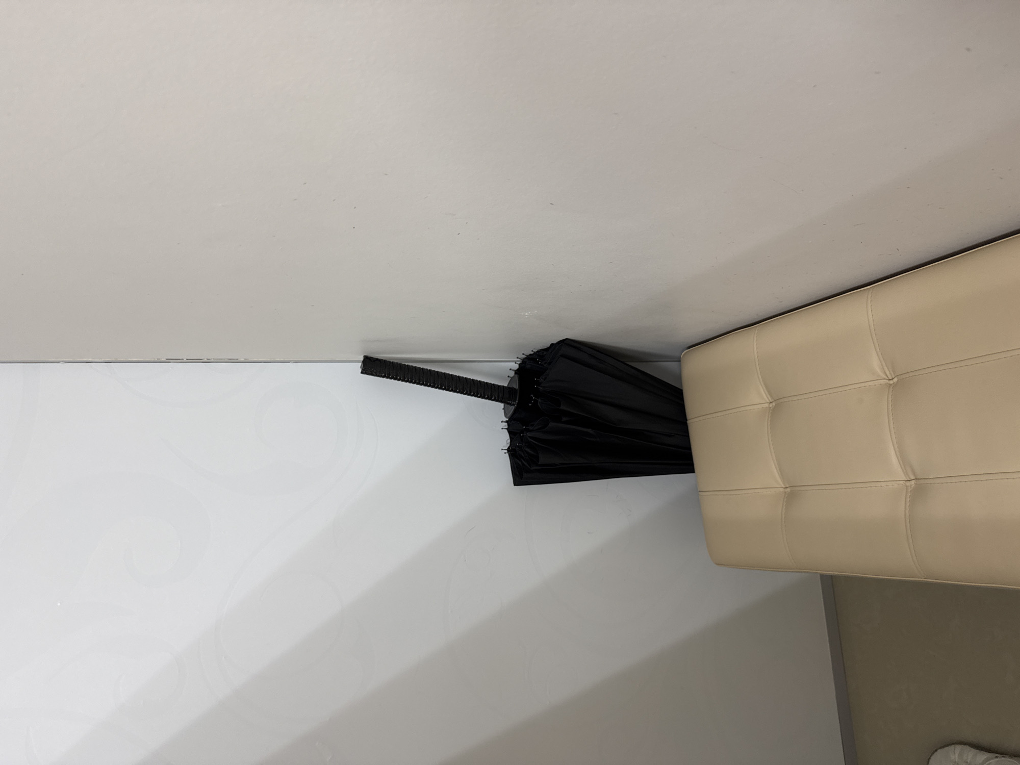 The image displays an indoor scene, likely a corner or narrow space. The main subject is a black, closed umbrella, positioned horizontally and tucked between a wall and a piece of furniture. On the left side of the image, there is a plain white wall with subtle, light-colored swirling decorative patterns. To the right, a light-colored (beige or cream) tufted piece of furniture, possibly an armrest of a sofa, a bench, or an ottoman, is visible, featuring square sections created by stitching. Above the umbrella and furniture, a uniform, light-colored ceiling or upper wall occupies the top portion of the frame. A small section of the floor, appearing light brownish-grey, is visible in the bottom right corner. No people are present, and no particular activity is taking place; the umbrella seems to be stored in this location. The lighting is consistent with an indoor setting. There is no visible text in the image.
