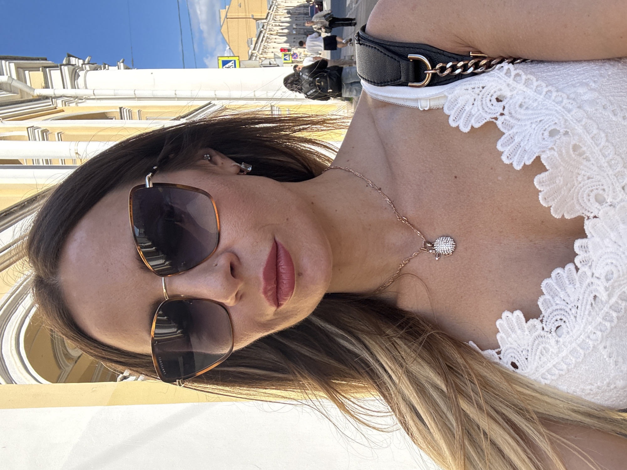 A woman is taking a selfie outdoors on a sunny day, with a street and buildings in the background. She is wearing large, tortoiseshell-patterned sunglasses, a white lace-trimmed top, and a delicate rose gold necklace with a round, diamond-encrusted pendant. Her brown hair is long and styled loosely. Behind her, a yellow building with arched windows and white columns is visible, typical of St. Petersburg's architectural style. In the background, several people are walking on a sidewalk, and a traffic light and street signs indicate an urban environment. The image is taken from the chest up, capturing the woman’s face and upper body.