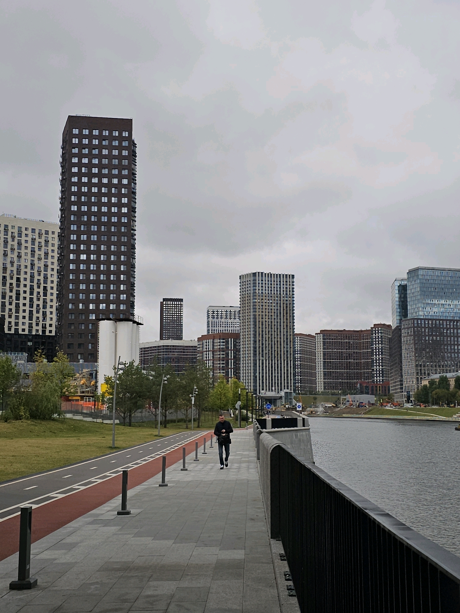 The image depicts a modern urban landscape with a view of the Moskva River. In the foreground, a man in a black jacket and pants walks along a paved walkway bordering a red bicycle lane. The walkway is lined with dark grey bollards and a black metal fence separates it from the river on the right. Across the river, a cluster of contemporary high-rise buildings forms the city skyline. These buildings vary in design and color, with some featuring dark facades and numerous windows, while others have lighter, multi-colored exteriors. Greenery, including trees and a grassy area, is visible on the left side of the walkway. The sky is overcast and grey, suggesting a cloudy day. The architecture and the presence of the Moskva River strongly suggest this location is in Moscow, Russia, likely in a newly developed or redeveloped area along the riverfront.
