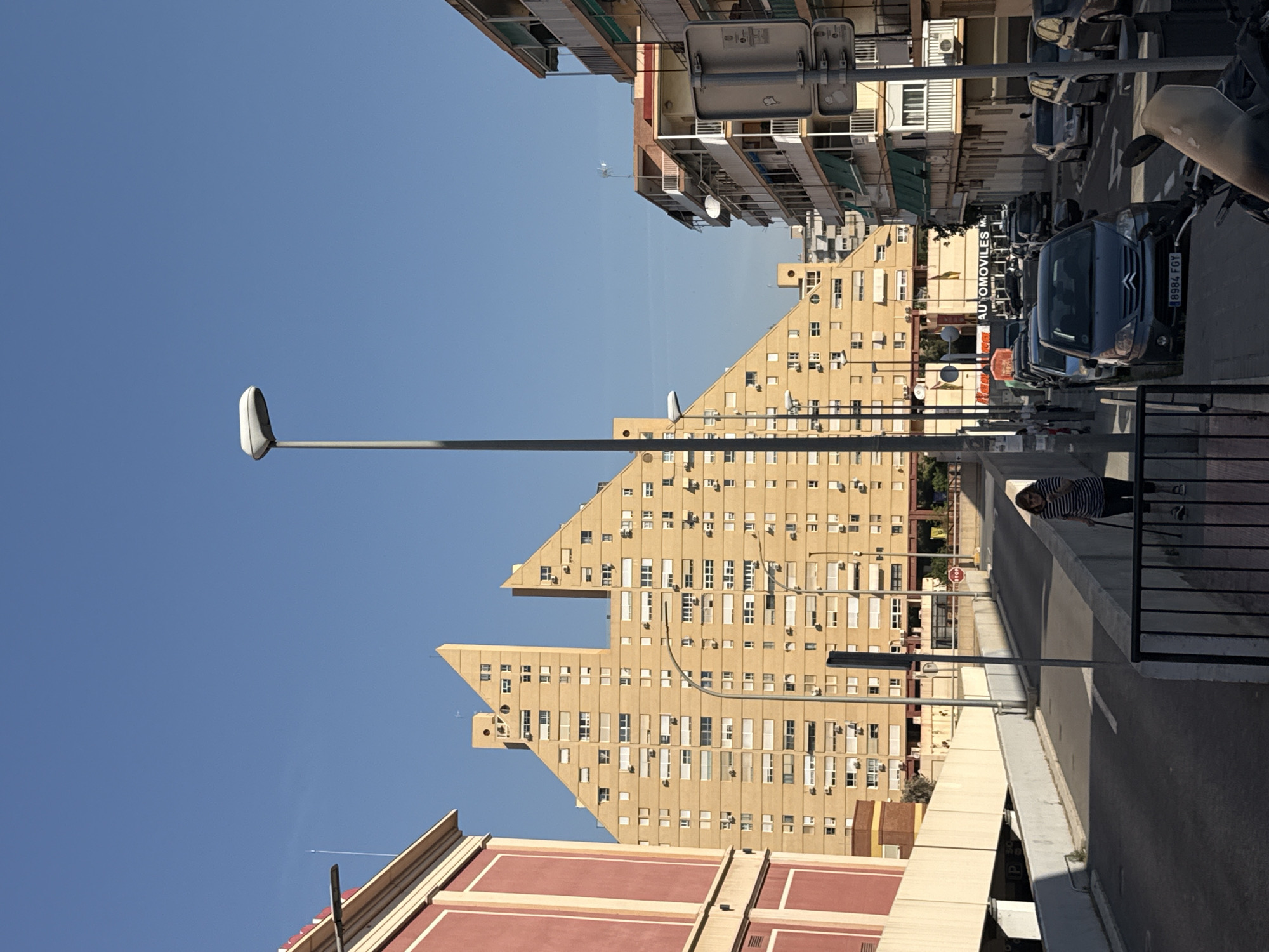 The image shows a city street in Unknown, Unknown, featuring tall, triangular buildings of beige concrete with rows of windows. A blue sky is visible above. A streetlamp stands tall on the left side of the image. Cars are parked along the street, with a grey Citroen visible on the right side. A person stands behind a railing on a raised walkway that runs along the right side of the street. A red and beige building is visible near the bottom of the image. Street signs and commercial signs are also present.