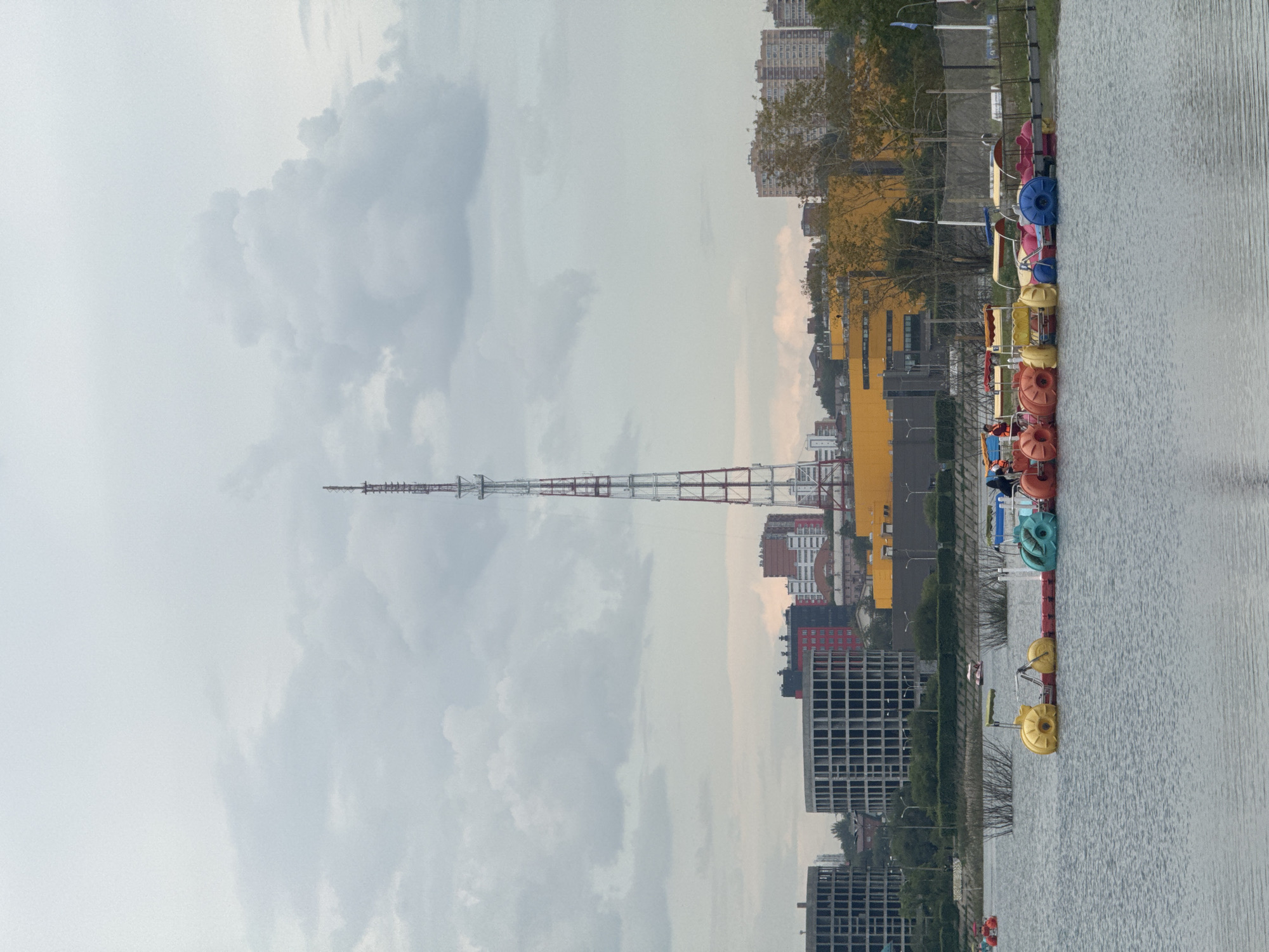 The image depicts a serene lakefront scene in Irkutsk, Russia, under an overcast sky. In the foreground, a fleet of colorful paddle boats are docked along a pier, some with passengers on board. The paddle boats are a mix of yellow, blue, red, and orange, with some featuring canopies. Beyond the lake, the cityscape of Irkutsk unfolds, characterized by a mix of modern architecture and infrastructure. Dominating the skyline is a tall, red and white radio tower, a prominent landmark in the city. Several multi-story buildings, including residential complexes and what appear to be commercial structures, are visible. The presence of lush green trees and bushes lining the embankment adds a touch of nature to the urban landscape. The overall atmosphere is calm and tranquil, suggesting a leisurely day at the lake.