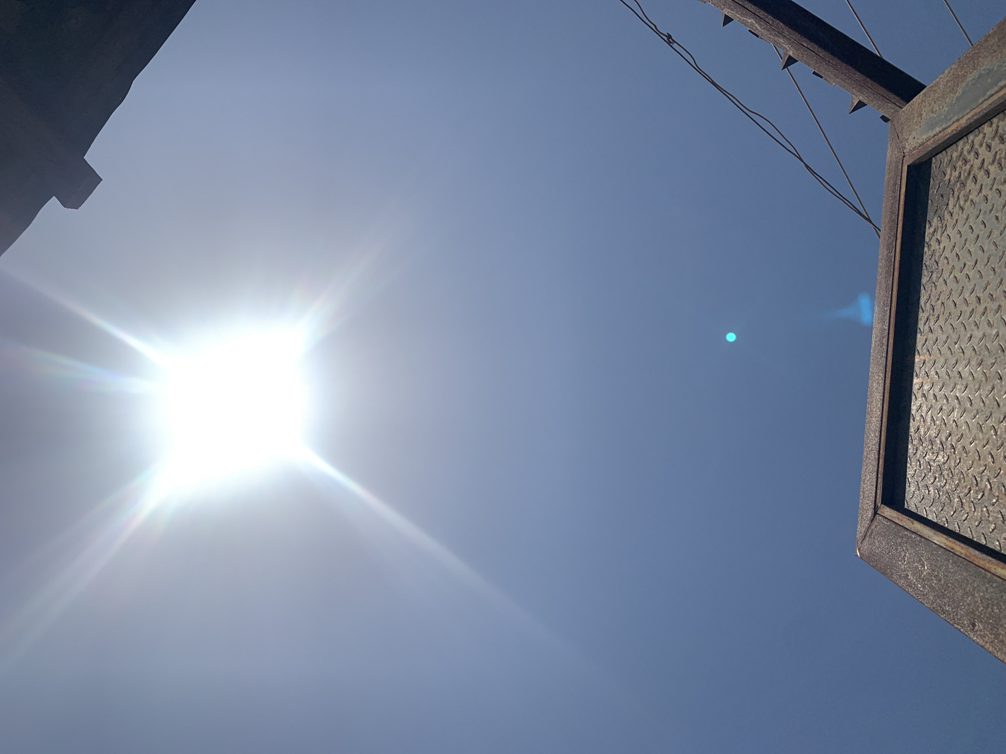 The image, taken from a low angle, features a bright sun against a blue sky. The top left corner shows part of a dark structure, possibly a building. In the top right, there's a metal frame with a textured surface, likely part of a gate or fence. Wires are visible extending from the frame. The location is purportedly Rafin Par, Nigeria, but the image alone does not provide explicit confirmation of this location. There are no people visible in the frame.