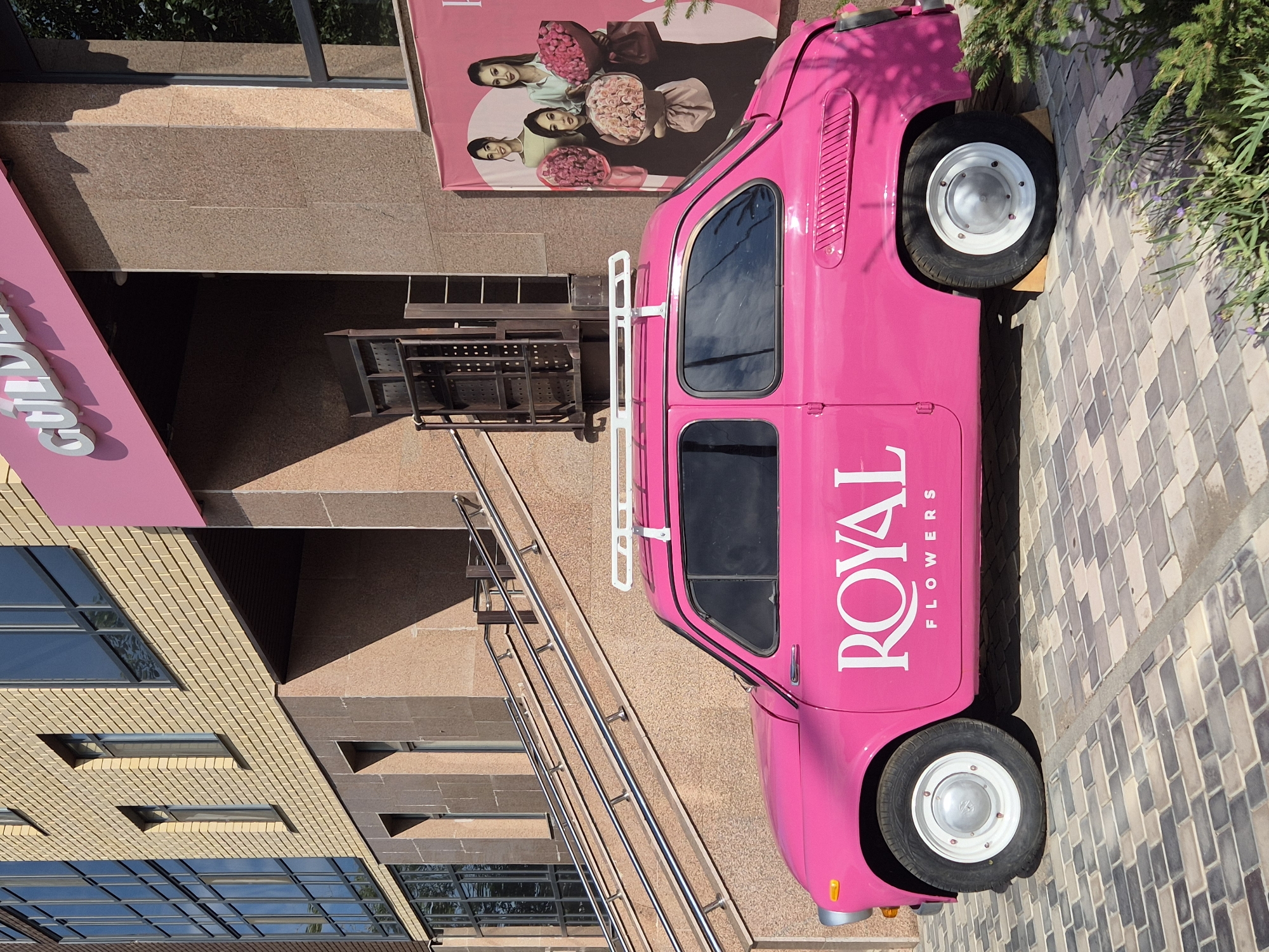 A bright pink vintage car, possibly a ZAZ-965 Zaporozhets, is parked on a cobblestone sidewalk in front of a modern building. The car has white lettering on its side that reads "ROYAL FLOWERS". A white roof rack is mounted on top of the car. The building features light brown brickwork and large windows. To the right of the car, a large pink banner displays images of four women holding bouquets of flowers. The scene suggests a commercial establishment, likely a flower shop, in an urban setting. The image was taken in Kokshetau, Kazakhstan.