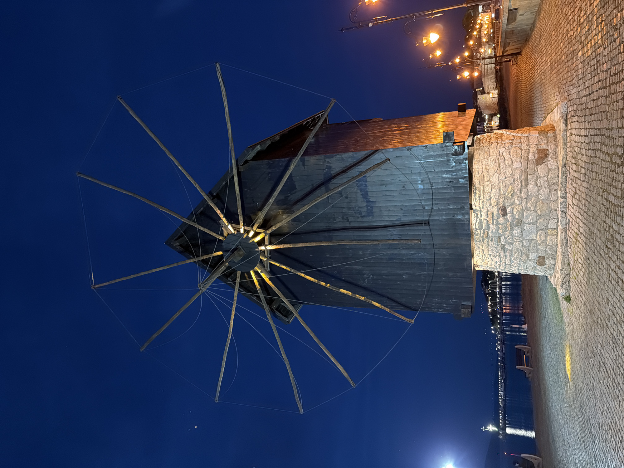 The image shows a windmill, a landmark in the city of Nesebar, Bulgaria, at night. The windmill has a dark wooden structure and multiple sails. It stands on a stone base. The background includes a waterfront area with lights reflecting on the water and a paved walkway with street lamps. The sky is dark blue.