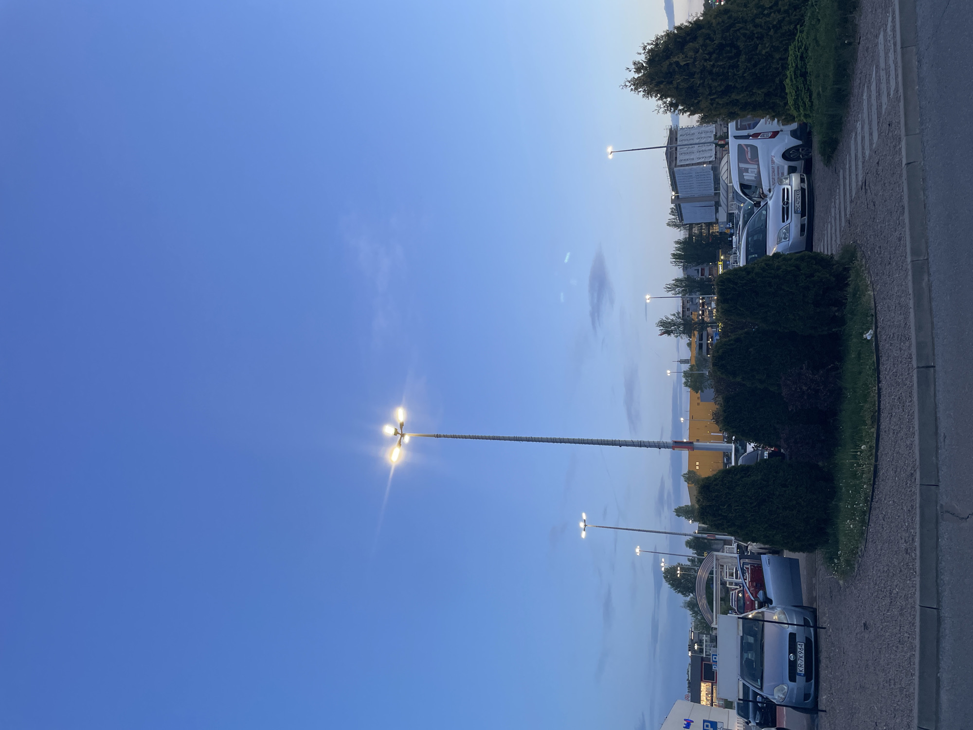 The image depicts a parking lot scene at dusk. Cars are parked along the right side of the frame next to a sidewalk and landscaping. Tall street lights illuminate the area. The sky is a gradient of blue. The city is unspecified.