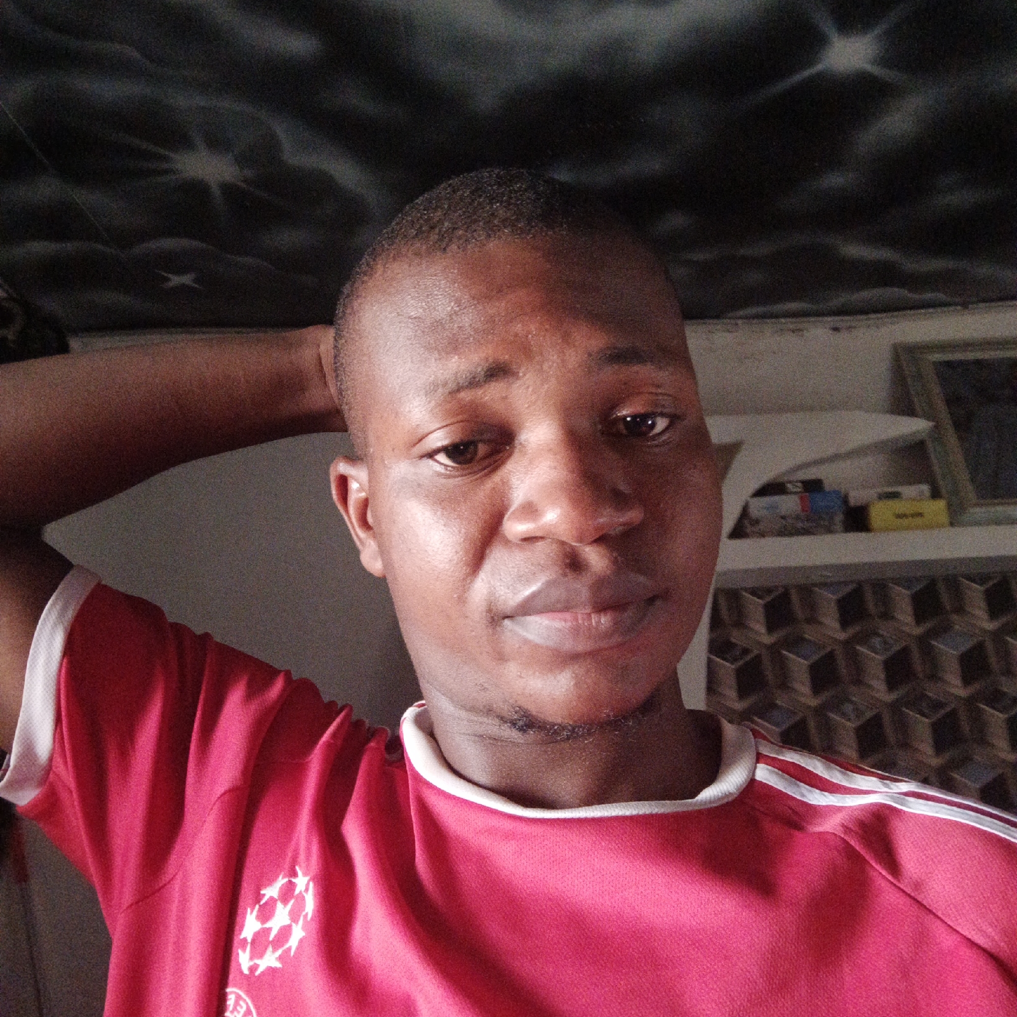 A medium, eye-level shot shows a dark-skinned man wearing a red shirt with white stripes, with his hand behind his head. He is likely indoors. There is shelving and wallpaper behind him. The city location is not discernible from the image.