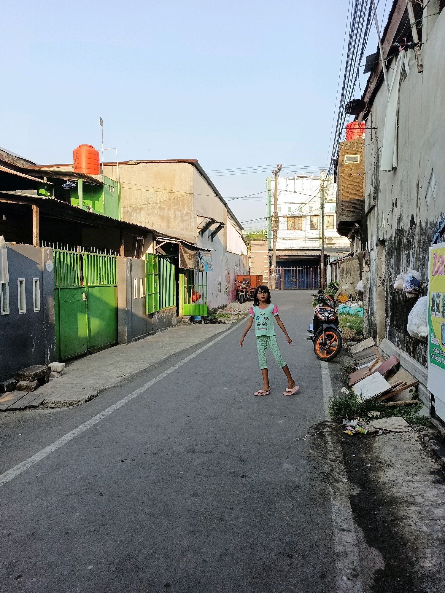 In Makassar, Indonesia, on a clear, sunny day, a young girl stands in the middle of a narrow, asphalted street. She is wearing a light green t-shirt with a graphic print and matching light green patterned pants, and pink flip-flops. Her arms are outstretched to the sides, and she appears to be posing or playing.
The street is lined with buildings of varying styles and conditions. On the left, there are structures with corrugated metal roofs and green painted fences and walls, suggesting a residential area. An orange water tank is visible on top of one of the buildings.
On the right side of the street, the buildings appear older and in a state of disrepair, with weathered walls and exposed materials. Various items, including discarded wood, debris, and plastic bags, are piled along the roadside. A motorcycle with an orange and black color scheme is parked on the right side of the street.
The overall impression is of a typical urban street scene in a developing area, with a mix of residential and commercial structures, and evidence of everyday life, including children playing outdoors and signs of urban living. The sky is a bright, clear blue, indicating good weather.