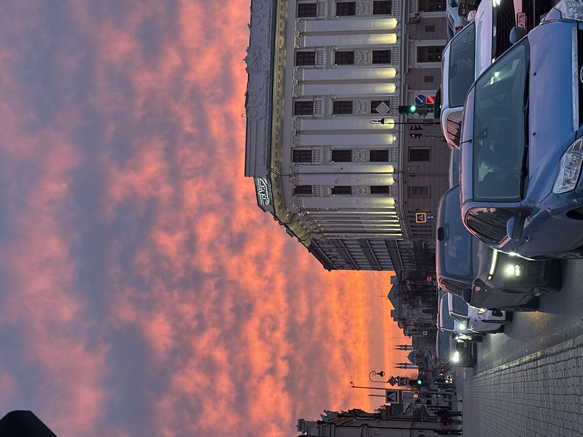 The image captures a street scene in Kazan, Russia, at dusk or dawn, indicated by the orange and purple hues of the sky filled with clouds. A row of cars, mostly facing forward, are parked along a cobblestone or paved street, occupying the right side of the frame. The first car in view is blue, followed by a mix of dark-colored and white vehicles.
On the left, a large, ornate building with many windows and illuminated columns dominates the scene. Traffic lights are visible, showing green, indicating a go signal. Further down the street, a glimpse of another building and street lamps can be seen. A few pedestrians are walking on the sidewalk. The building appears to be of significant architectural importance, suggesting it is a landmark in the city. The scene presents a typical urban setting with transportation, architecture, and the daily movement of people.