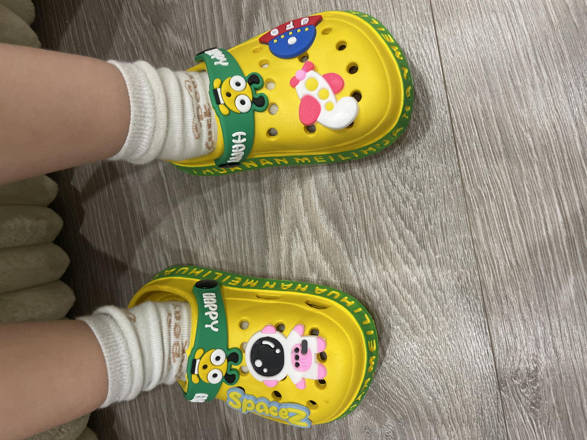 The image displays a close-up, top-down view of a person's feet, presumably a child's, wearing white socks and bright yellow clogs. The setting appears to be indoors, on a light grey wooden or laminate floor with distinct grain patterns. A light-colored, soft object, possibly a blanket or cushion, is partially visible on the left side of the frame.
The feet are clad in white, ribbed socks that are slightly crumpled around the ankles. Both feet are resting on the floor, one slightly above the other.
The clogs are a vibrant yellow, featuring multiple ventilation holes across the top. They have green, adjustable heel straps. The straps on both clogs have a yellow cartoon character with large eyes, possibly a bear or a similar animal, and the word "HAPPY" embossed in white. The side edges of the yellow clogs are also green, with the text "HUANAN MEILIHUA" repeatedly embossed around the rim.
Each clog is adorned with space-themed decorative charms. The upper (left) clog features a blue and red spaceship or airplane and a white and pink rocket or plane with yellow polka dots. The lower (right) clog has a white and pink astronaut figure and a blue and yellow emblem that reads "Space Z".
No specific activity is taking place other than the wearing of the footwear. The time of day and any emotions are not discernible from the image.