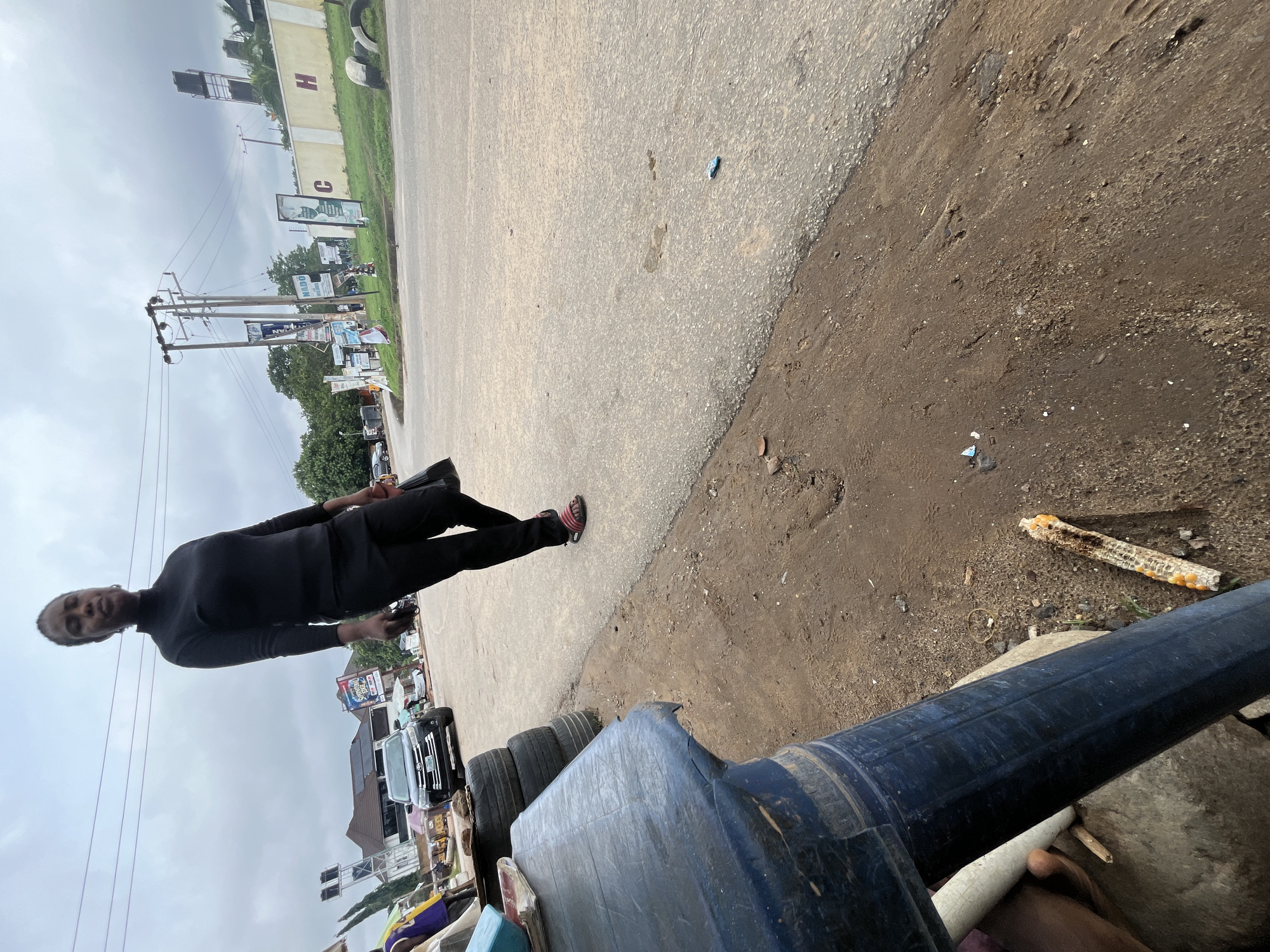 A full shot photograph taken from a low angle shows a Black woman walking on a paved road on an overcast day. She is wearing a black turtleneck shirt and black pants, with red sandals on her feet. She is holding a black bag in her right hand. To the left of the woman, a dark blue plastic table is in the foreground, with a stack of tires behind it. In the background, there is a black SUV, and several buildings and billboards. The scene appears to be a roadside in Abuja, Nigeria, with power lines and streetlights visible. The ground is dirt and asphalt, with some debris scattered on the roadside.