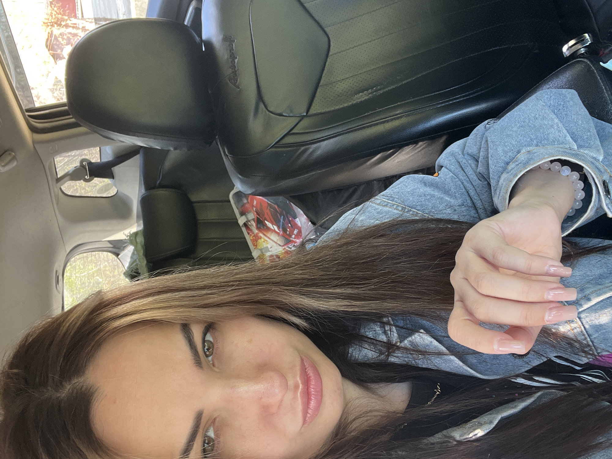 A selfie of a young woman with long brown hair and fair skin, wearing a light denim jacket and a beaded bracelet, taken inside a vehicle. The interior of the vehicle includes black leather seats and a visible window showing trees and exterior light. The location is Unknown, Unknown.