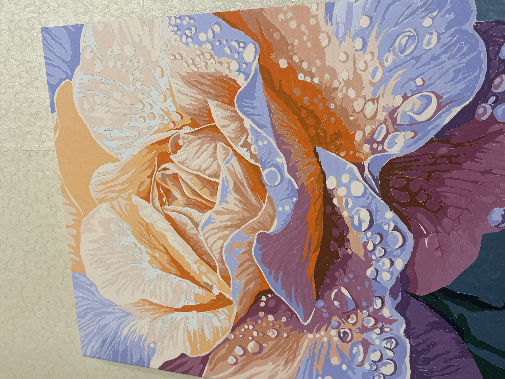 This is a close-up, eye-level, studio shot of a painting of a rose with water droplets on its petals. The painting is laid flat on a textured, light-colored surface, likely a table or wall, which appears to be a light beige with a subtle floral pattern. The painting itself is rendered in a variety of pastel colors, including shades of orange, peach, lavender, purple, and some darker greens and browns. The rose is the sole subject, depicted with soft, layered petals that appear to be made of fabric or paper, given the stylized rendering. Numerous small, circular shapes with white highlights, representing water droplets, are scattered across the petals, particularly on the lower half of the composition. Some of these droplets have a slight blue or pinkish hue. The painting has visible brushstrokes and areas where numbers are printed, indicating it is a paint-by-numbers artwork. The lighting is even, casting no distinct shadows, suggesting a controlled studio environment or indirect natural light. The focus is sharp on the painting, with the background slightly out of focus.