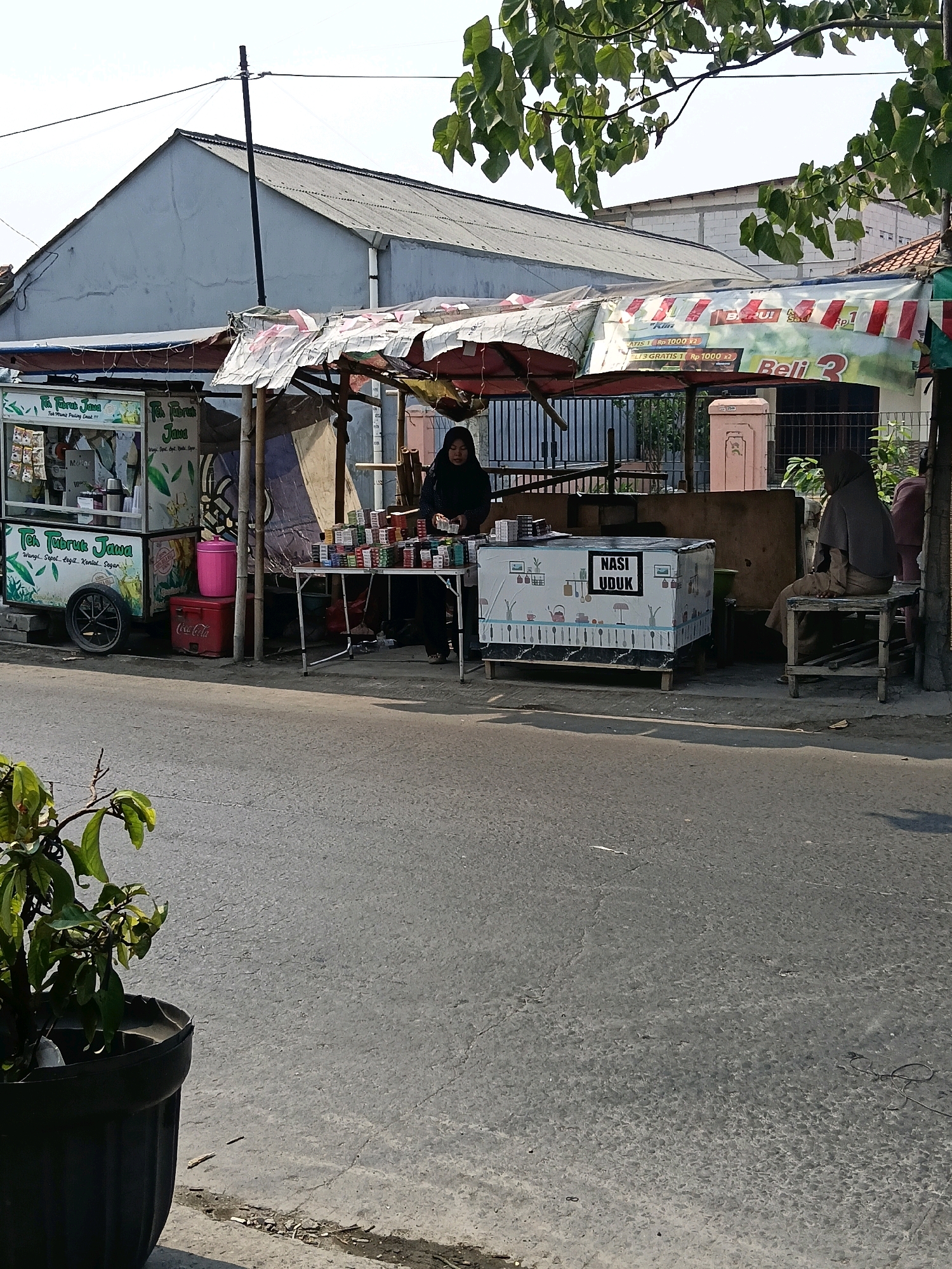 The image captures a street scene in Belimbing, Indonesia, likely during daytime with clear skies and bright sunlight casting shadows. In the foreground, a potted plant with green leaves is partially visible on the left, placed on a concrete surface. A paved road occupies the majority of the lower frame.
The mid-ground features a roadside stall with various offerings. A small cart, possibly a mobile food or drink vendor, is positioned on the left. It displays signage for "Teh Tarik Jawa" (Javanese Milk Tea) and has products arranged on its shelves. A pink cooler with a Coca-Cola logo is visible next to the cart.
Further to the right, a woman wearing a dark headscarf and black clothing stands behind a foldable table laden with numerous packs of cigarettes. She appears to be attending to a customer or organizing the merchandise. Adjacent to this, a white rectangular structure, possibly a cooler or display unit, is wrapped with a decorative cloth featuring kitchen utensils and cutlery. A black sign on this unit reads "NASI UDUK" (Coconut Rice), indicating it's a food stall.
Another person, also wearing a headscarf and brown attire, is seated on a wooden stool to the right of the "Nasi Uduk" unit, observing the surroundings. Behind the stalls, a fence encloses an area with some vegetation and buildings. A banner with promotional text, including prices and offers in Indonesian, is suspended above the stalls. The overall environment suggests a typical street-side commercial setup in a residential or semi-urban area.