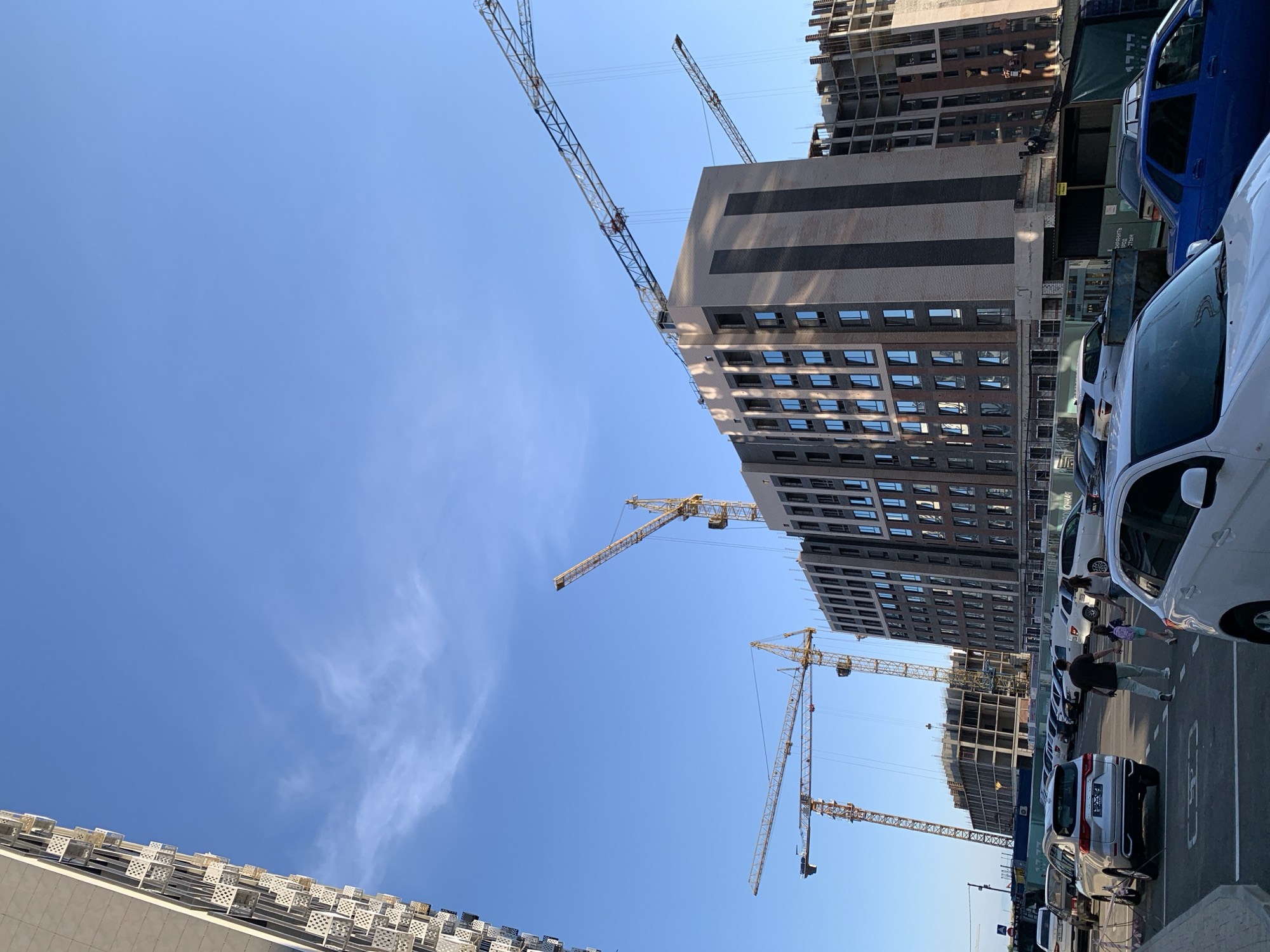 The image depicts a construction site in Stavropol, Russia, under a clear blue sky with a few wispy clouds. In the foreground, a parking lot is filled with various cars, including a silver SUV, a white sedan, and a blue sedan. A man and a young girl are walking across the parking lot.
Dominating the background are several tall buildings under construction. One building is nearing completion, featuring a modern facade with numerous windows. Multiple construction cranes, primarily yellow, are active at the site, with their booms extending high into the sky. A partially constructed building, appearing to be a concrete frame, is visible behind the more complete structure. To the left, a tall residential building, likely already occupied, displays a distinctive patterned facade. The overall scene suggests ongoing urban development.