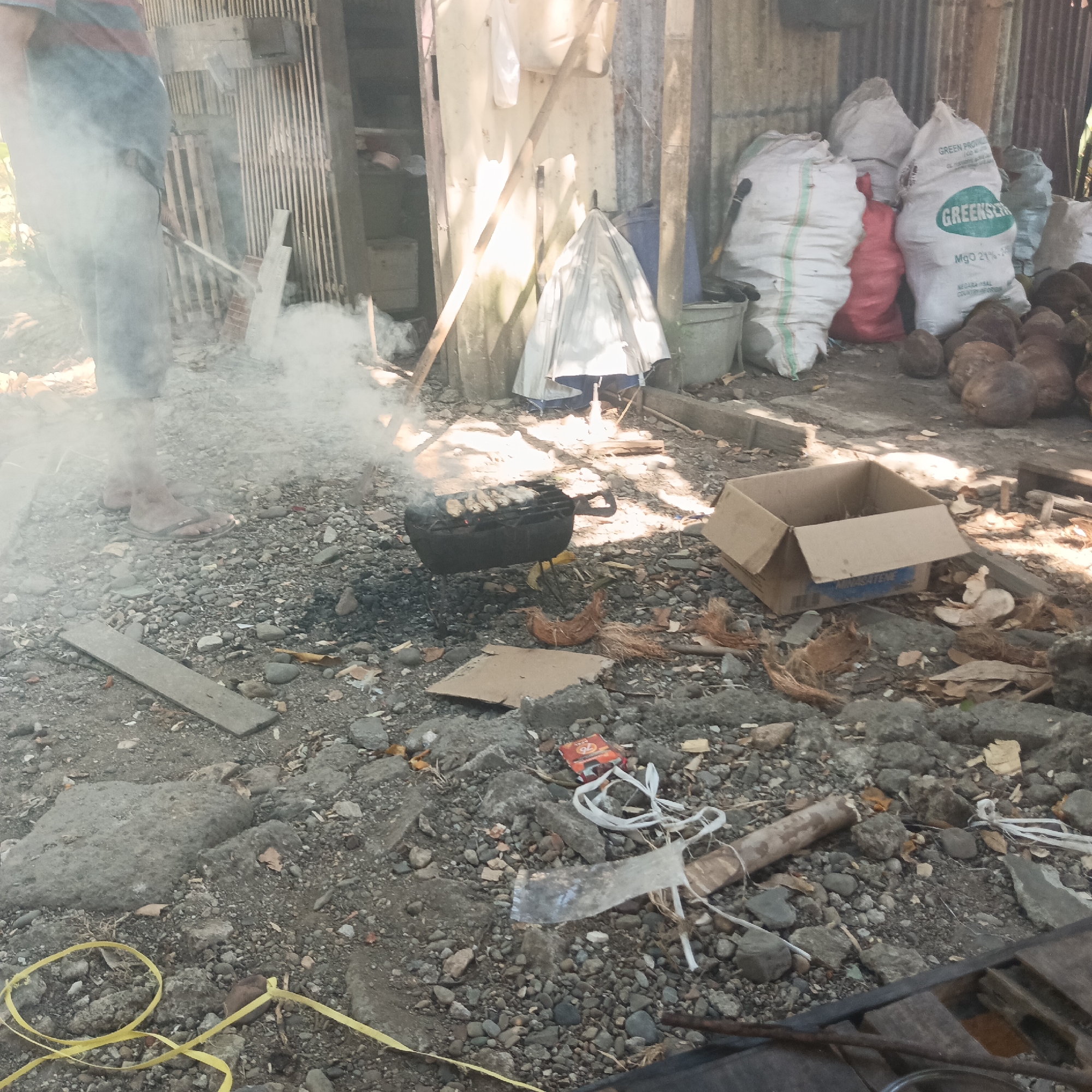 A person stands in a smoky outdoor area, their lower legs and feet visible, wearing flip-flops and dark pants. They are positioned near a black charcoal grill on which food is being cooked, with smoke rising from the grill. The ground is covered in gravel and rocks, with scattered debris including coconut husks, pieces of cardboard, and a yellow plastic ribbon. In the background, several large sacks are stacked against a corrugated metal wall, and other objects like buckets, a folded umbrella, and coconuts are visible. The scene appears to be an outdoor cooking area in a rustic or informal setting within the city of Tumampua, Indonesia.