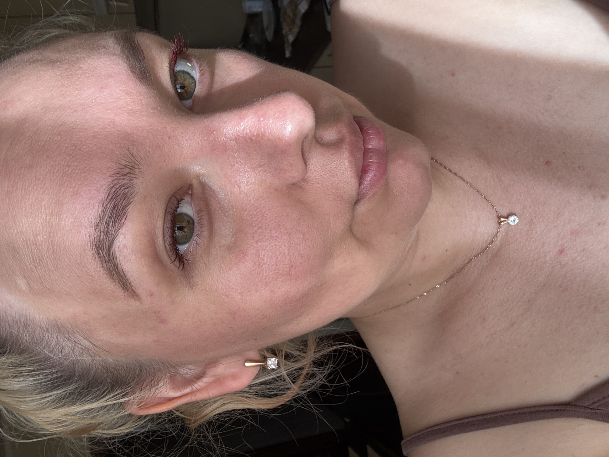 This is a close-up, eye-level, outdoor medium shot of a fair-skinned woman's face and upper chest. The woman has blonde hair pulled back, brown eyebrows, and green eyes. Her skin appears flushed, with some visible pores and redness. She is wearing a gold earring with a small square diamond, and a delicate gold necklace with a single diamond pendant. Her lips are slightly parted, and she is looking towards the camera. The background is dark and out of focus, suggesting an indoor setting or late evening. There are no specific elements in the image that can be used to determine the location in Chaikovsky, Russia.