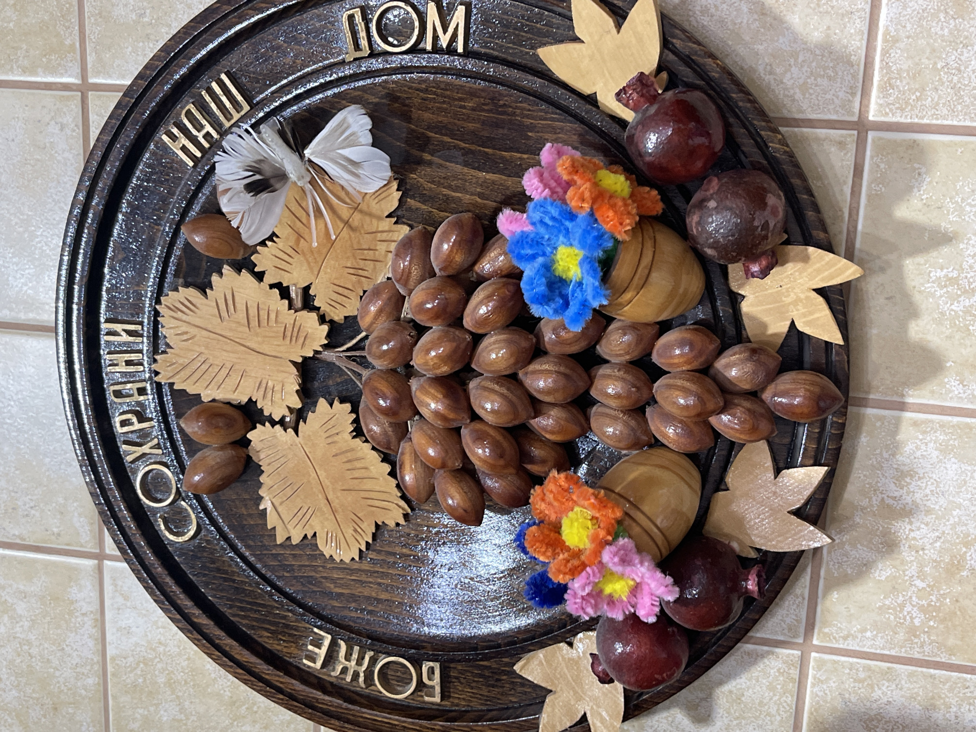 The image depicts a handcrafted, circular wooden platter. The platter is decorated with a cluster of brown, nut-like objects, wooden leaves, and small wooden vases filled with colorful, fuzzy flowers. Dark red, fruit-like objects are positioned around the platter's edge. The platter has a dark brown border inlaid with gold-colored Cyrillic lettering, partially obscured. The overall impression is that of a rustic, homemade decorative piece. The location is unspecified, but the image description references the city of Donskoye, Russia. No people are present in the image. The subjects are decorative items. The primary objects are the wooden platter, nuts, wooden leaves, wooden vases, fuzzy flowers, and fruit. The actions involve the arrangement of the decorative items on the platter. The environment is a tiled surface, likely a kitchen or bathroom.