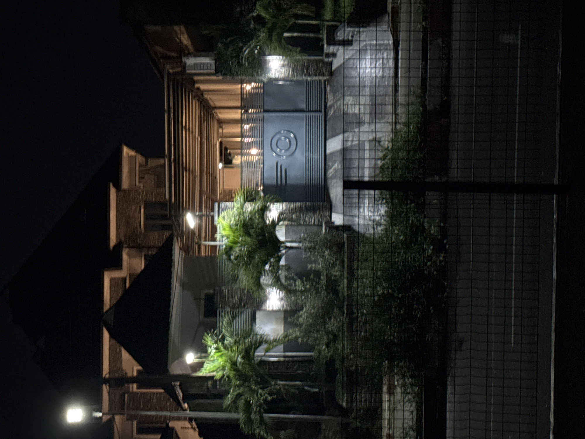 The image shows a dimly lit scene at night, likely in the city of Uyo, Nigeria. In the foreground, a black wire fence runs horizontally across the frame, with a dark road or pavement behind it. Beyond the fence, lush green foliage and palm trees are visible, illuminated by artificial lights. Behind the vegetation, a modern building with multiple levels and a dark, pyramidal roof is partially visible. The building appears to have brick and concrete facade elements. A prominent feature is a large, dark metal gate with a circular logo embossed on it, situated in front of the building. The gate is flanked by stone pillars and is set against a backdrop of covered walkways or pergolas supported by horizontal beams. Several lights are visible within the covered area. An outdoor air conditioning unit is mounted on the exterior wall of the building to the right. The overall impression is of a well-maintained property entrance at night.