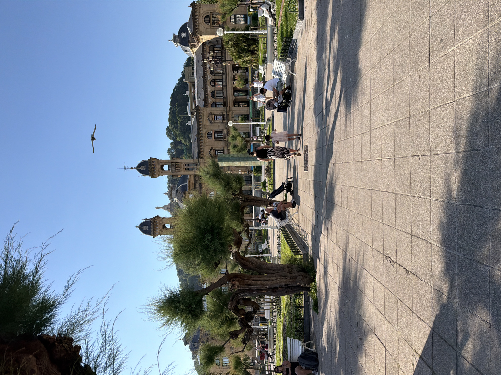 This photograph captures a scene on a paved walkway in San Sebastián, Spain. The walkway is constructed of square gray tiles and is partially shaded by trees. In the distance is a grand, multi-storied building with a distinct architectural style. The building features arched windows, detailed cornices, and domed towers. It is likely the San Sebastián City Hall, previously the Gran Casino, an iconic structure in the city.
Several people occupy the walkway. Some are sitting on benches, while others are walking or standing. A family is gathered around a stroller. The trees lining the walkway are interspersed with ornamental bushes and a short metal fence. In the sky, a bird is in flight.