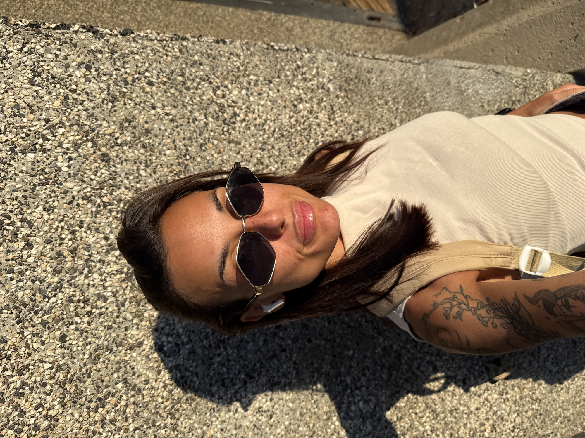 A young woman with dark hair, sunglasses, and earbuds stands against a textured wall. She is wearing a light-colored, ribbed tank top and a tan backpack strap is visible over her shoulder. The lighting suggests it is late afternoon or early evening, casting a long shadow from her left. The background wall is made of small, irregular stones. Her left arm, visible in the foreground, is adorned with tattoos. The image was captured in the city of Pleven, Bulgaria.