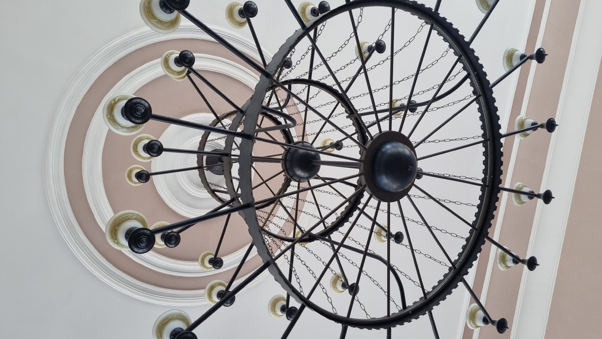 An indoor, low-angle shot captures an ornate, multi-tiered chandelier hanging from a ceiling with decorative molding. The chandelier is constructed of dark metal, featuring multiple circular frames connected by spokes and chains. It holds numerous light fixtures with creamy, opaque glass shades and black accents. The ceiling is painted white with a band of pale pink and intricate white molding, creating a layered effect. The focus is on the chandelier and the ceiling above it. The image does not provide any information about the specific location within Mukachevo, Ukraine, or the presence of any people.