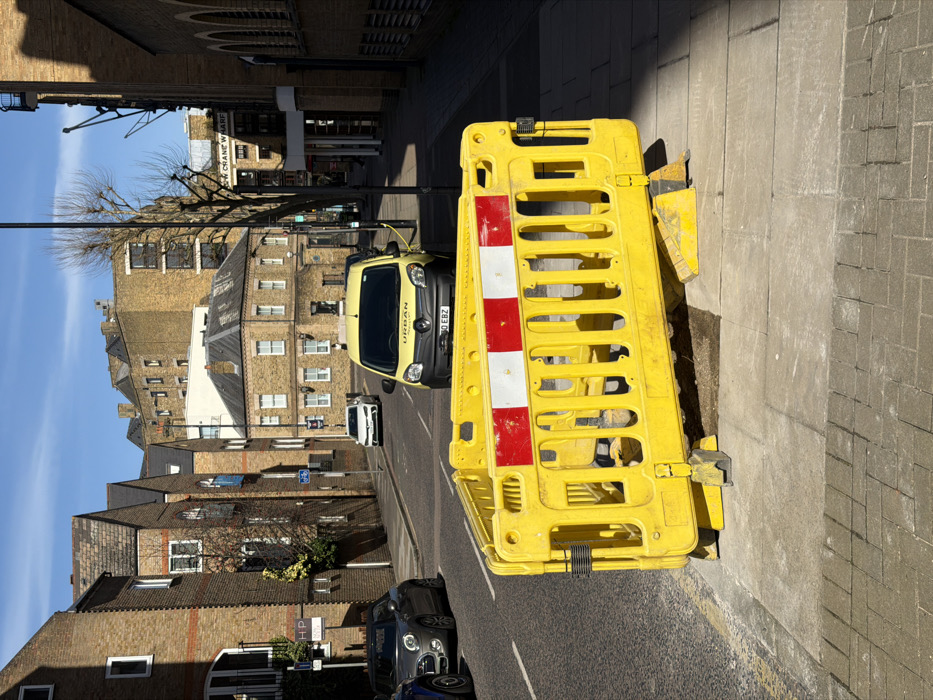The image depicts a sunlit urban street scene in Wapping, London, United Kingdom, specifically identifying Bow Crane Wharf through a visible street sign. In the immediate foreground, a bright yellow plastic temporary barrier, approximately one meter in height, stands on the pavement. This barrier features three prominent horizontal red and white reflective stripes across its upper section and rests on dark, possibly recycled rubber, feet. It partially encloses an area of disturbed light brown earth or an open utility trench on the grey paved sidewalk.
The street, constructed of asphalt, curves gently to the left and is lined with parked vehicles. A grey Mini Cooper is parked on the left side of the street, followed by a yellow Renault Kangoo van, branded "URBAN" on its bonnet and displaying the UK license plate "BO EBZ", positioned facing the viewer. A white car is visible parked further down the street behind the yellow van. On the left side of the street, a "P HP" parking sign is attached to a lamppost.
The background is dominated by multi-story brick buildings, characteristic of London architecture, with varying rooflines and window styles, including some arched windows. A visible street sign on the right identifies "BOW CRANE WHARF". Several bare trees are interspersed among the buildings and along the street. The sky is clear and blue. No individuals are visible in the scene.