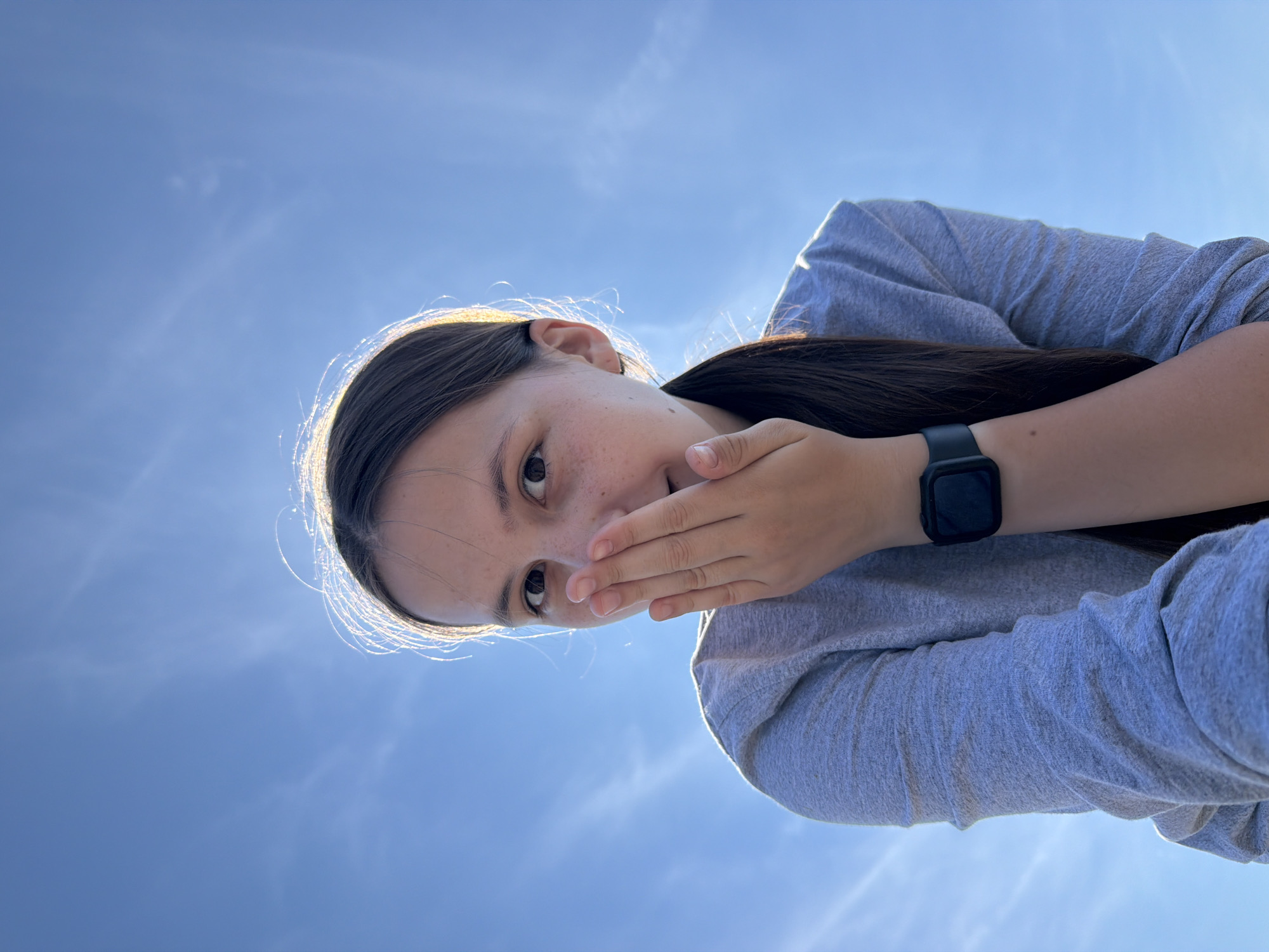 A young girl with dark hair and freckles is captured from a low angle, looking up at a bright blue sky with wispy clouds. She is wearing a grey long-sleeved shirt and a black smartwatch. Her right hand is covering her mouth, and she appears to be smiling or trying to suppress a laugh. The sunlight is catching the edges of her hair, creating a halo effect. The background is entirely the sky, indicating an outdoor setting. The image does not contain any information that places the location in the city of Stepanovo, Russia.