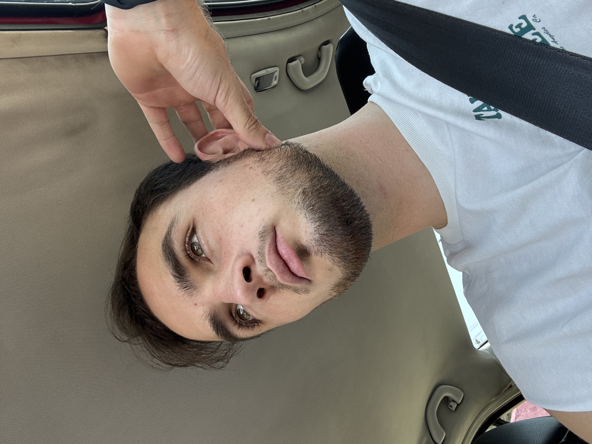A young man is captured in a close-up, angled portrait, appearing to be taking a selfie or having his picture taken inside a car. His head is tilted, and his left hand is raised, with fingers gently touching his left ear. He has dark hair, prominent brown eyes, a short beard or stubble, and light pink lips. A small reddish mark is visible on his forehead/cheek.
He is wearing a white t-shirt with partial dark green text visible, showing "E" and "LA". A dark seatbelt crosses his chest and shoulder. The background reveals the car's interior, including a light grey or beige headliner and part of a car door with a handle. The lighting suggests it is daytime. The man looks directly at the camera with a neutral to slightly pouting expression. This scene is located in San Bernardino Tlaxcalancingo, Mexico.