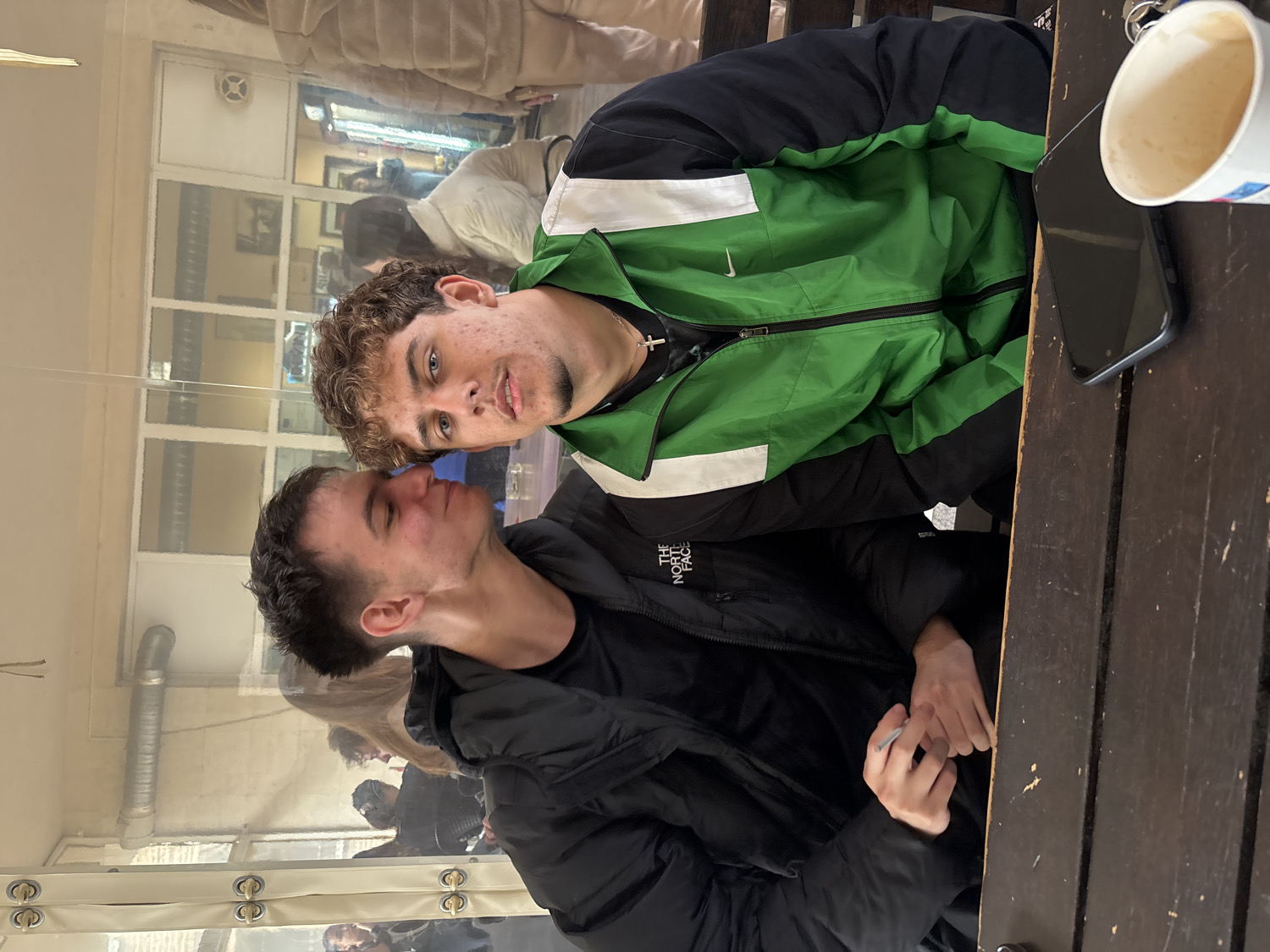 Two young men are seated at a wooden table, posing for a photograph in what appears to be a covered outdoor seating area or a cafe. The image is oriented vertically, showing the subjects from the chest up.
The man on the right (with curly brown hair and light stubble) wears a green and black jacket with a white stripe and a visible "Nike" logo. A silver cross pendant hangs around his neck. He looks directly towards the camera with a neutral expression.
The man on the left (with short, dark hair and clean-shaven) wears a black puffer jacket with a "The North Face" logo visible on his left shoulder. He is looking slightly upwards and to his right, towards the other man, with a calm expression. His hands are clasped on the table, and he appears to be holding a small, white, cylindrical object, possibly a cigarette or a similar item, between his fingers.
On the wooden table, next to the man in the green jacket, rests a black smartphone and a white paper cup containing a light brown beverage, likely coffee.
The background, partially obscured by reflections, shows interior elements such as white walls, a white ventilation grate, and what appears to be a glass or transparent plastic enclosure, suggesting a sheltered dining or social space. Reflections of other people and a street scene can be vaguely discerned in the background. The overall lighting suggests it is daytime. The scene is set in Plovdiv, Bulgaria.