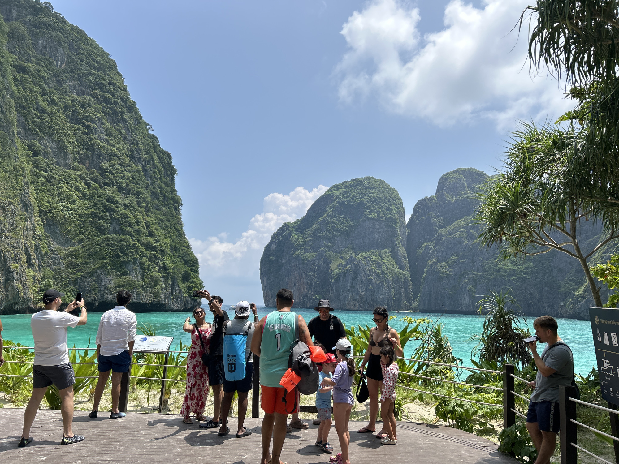 In Ao Nang, Thailand, a group of tourists stands on a wooden boardwalk overlooking a vibrant turquoise bay surrounded by lush green limestone cliffs. The sky is a bright blue with scattered white clouds. The people, including adults and children, are dressed in casual summer clothing, with some holding phones and cameras, suggesting they are taking pictures or videos. One person is holding a blue dry bag with "Ocean Pack 15" printed on it. The boardwalk has a white rope railing with wooden posts, and tropical foliage, including palm-like plants, lines the edge. The water is clear, revealing the sandy bottom in some areas, and a few small boats are visible in the distance. The scene captures a moment of leisure and sightseeing in a picturesque coastal setting.