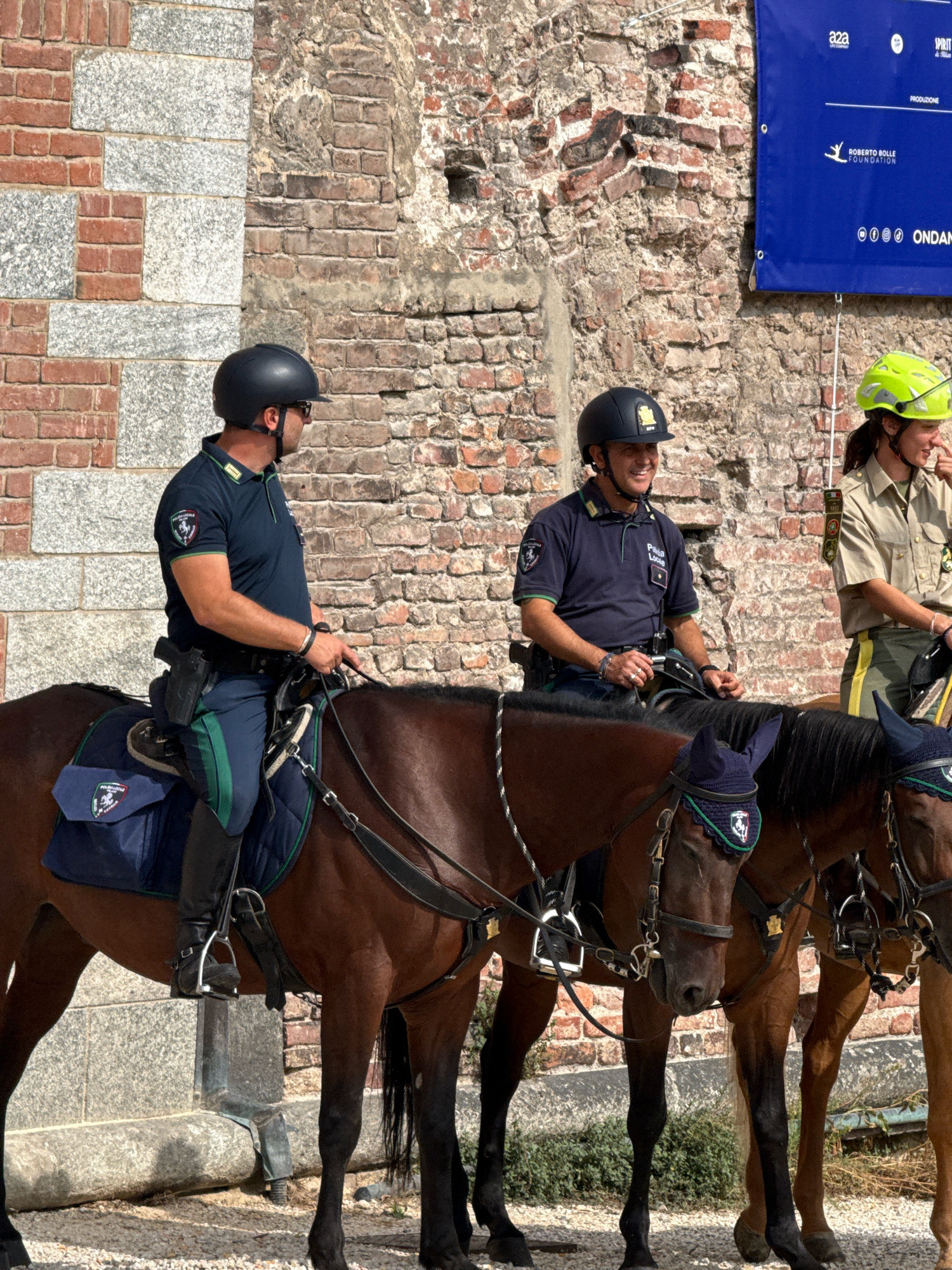 Two mounted police officers, one wearing a dark blue uniform with green trim and a black helmet, and another wearing a khaki uniform with a bright green helmet, are positioned in front of a brick wall. The officer in the dark blue uniform is on the left, smiling, with a handgun holstered on his right hip. He wears black boots and has a saddlebag with a police insignia on his horse. The officer in the khaki uniform is on the right, with a hand to her face, appearing to be speaking. Both horses are fitted with bridles and saddles. Behind them, a blue banner is partially visible, displaying text including "ROBERTO BOLLE FOUNDATION" and "ONDANOMIE". The background is a weathered brick wall. The scene appears to be outdoors, with sunlight casting shadows. This location is likely in Milan, Italy, given the prominent display of the Roberto Bolle Foundation, a Milan-based organization.