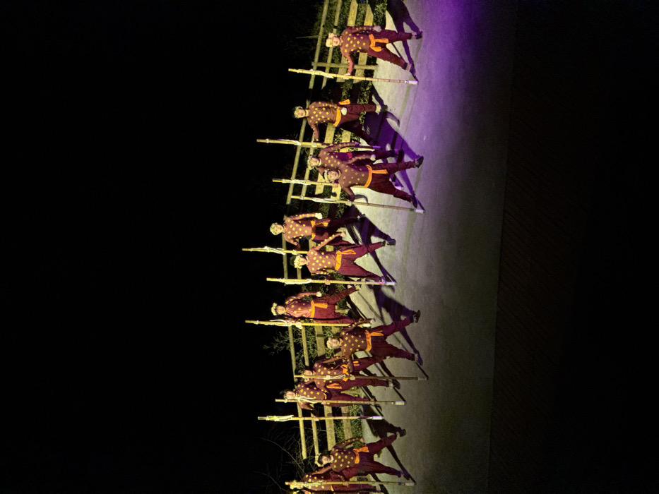 An outdoor night performance is underway in Wulong District, China, featuring approximately 11-12 male performers arranged in rows on a light-colored paved surface. Each performer is dressed in a dark red or maroon long-sleeved top and matching pants, adorned with golden-yellow star-like patterns, and a prominent orange sash around the waist. They are holding long, light-colored wooden or bamboo poles vertically with both hands, adopting a static, synchronized pose. Bright artificial lighting illuminates the performers from above, casting distinct shadows on the ground, which also displays a faint purple hue on the right side. Behind the performers, a light-colored wooden fence separates the stage from a dark, indistinct vegetated background, likely trees or bushes. The sky above is completely black, indicative of nighttime.