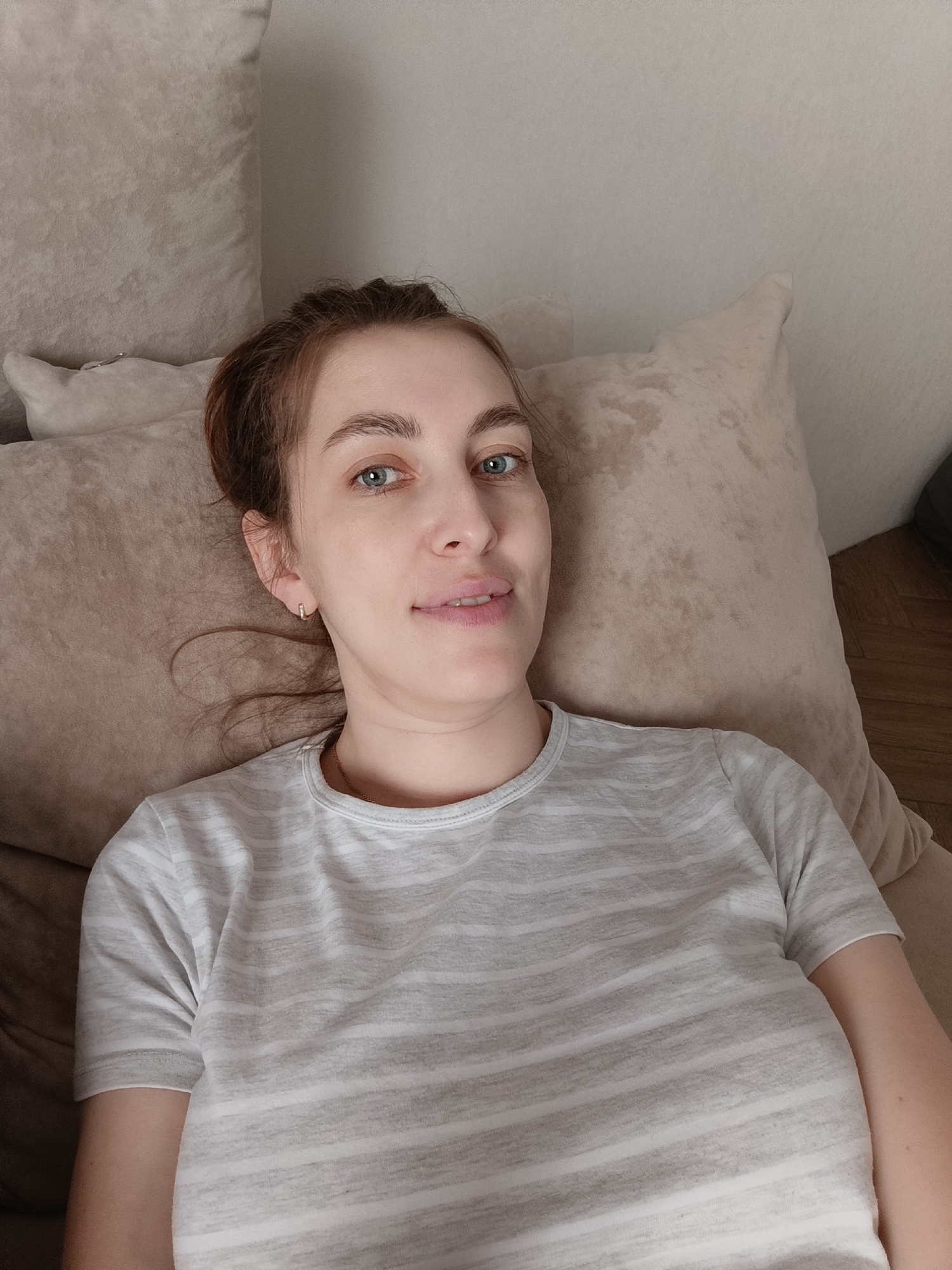 A woman is lying down on a beige couch or bed, facing the camera. She has light skin, light blue eyes, and her brown hair is pulled back in a bun. She is wearing a grey and white striped t-shirt and small gold hoop earrings. The background includes beige pillows and a portion of a wooden floor. There is no information present in the image to suggest that it was taken in the city of Chaikovsky, Russia.