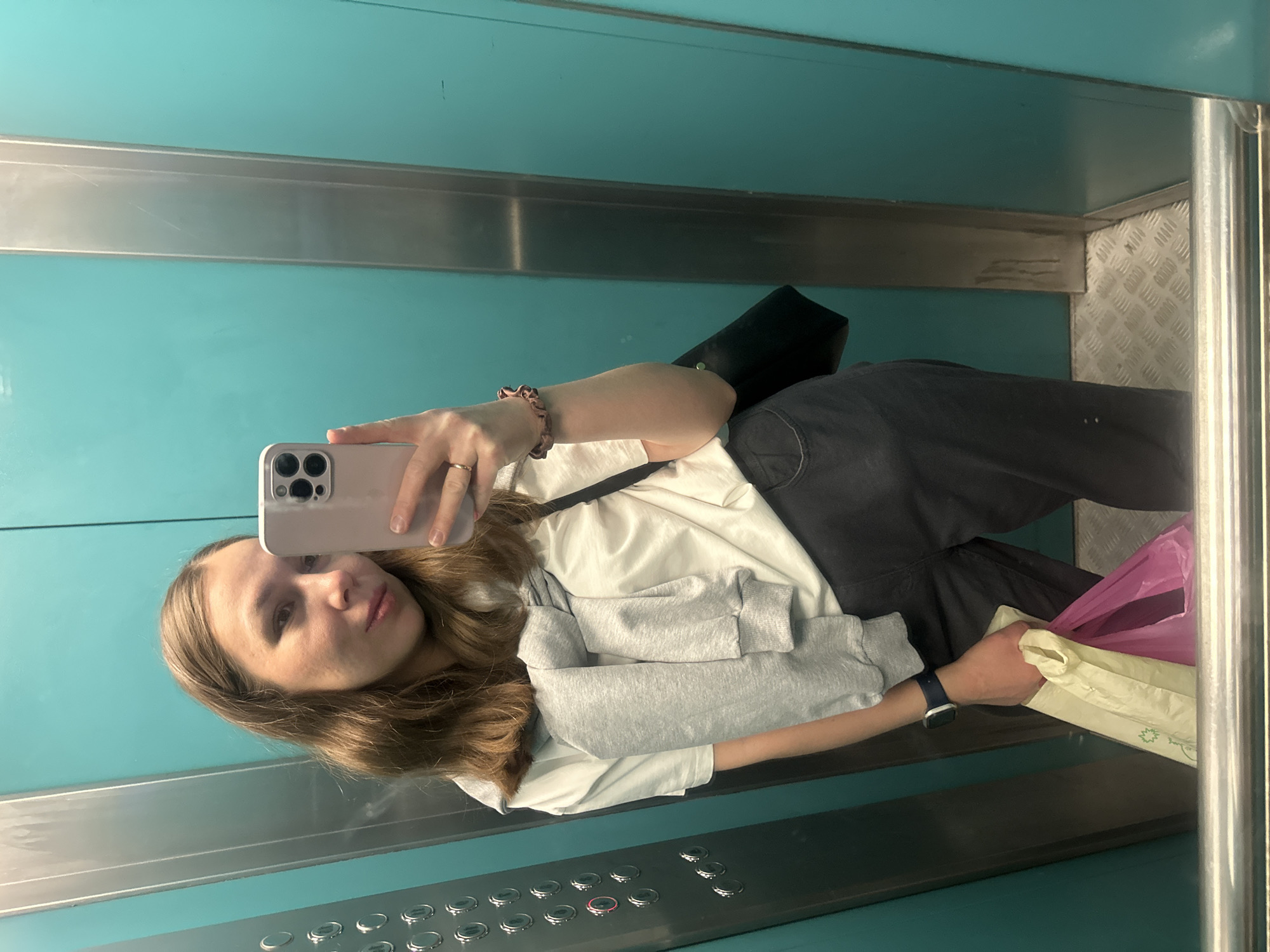 A woman is taking a selfie in an elevator. The walls of the elevator are teal-colored, and the floor appears to be a metallic, non-slip surface. The elevator's control panel is visible, featuring several circular buttons. The woman holds a phone in her left hand to capture the image. She's wearing a white t-shirt, a grey sweatshirt, and black pants. She is also wearing an Apple Watch on her left wrist. She has a ring on her right ring finger. A black purse hangs on her shoulder. She is holding a light yellow bag and a pink bag in her right hand. The location is possibly Tomilino, Russia.
