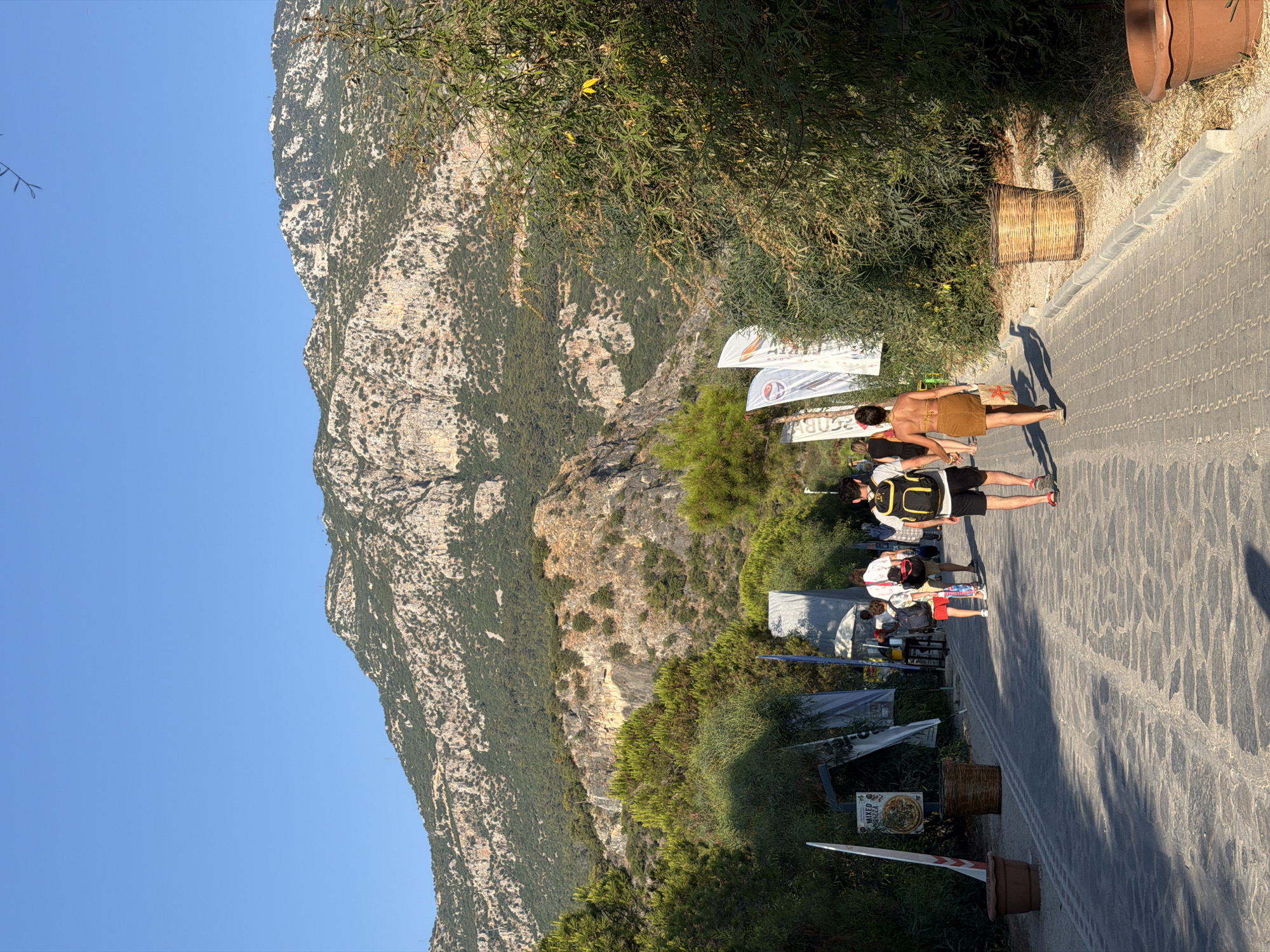 The image depicts a sunny day in Fethiye, Turkey, with a mountainous backdrop. A paved walkway, constructed with irregular stone tiles, leads away from the viewer. Several people are walking on the path, some carrying backpacks, suggesting a leisure or travel context. The path is lined with lush green vegetation on both sides, including trees and bushes. Flags with various logos and text are planted along the walkway, indicating a possible event or commercial area. Two large, woven baskets and terracotta pots are placed along the edge of the path, adding decorative elements. In the distance, a rugged mountain range dominates the horizon, its slopes covered with sparse vegetation and rocky outcrops. The sky is clear and blue.
