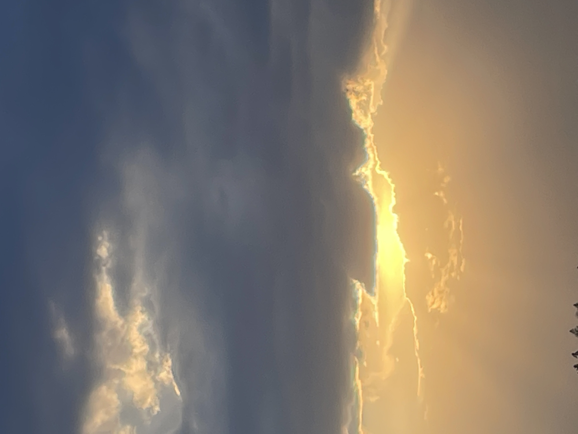 The image shows a sky with clouds. The left side of the image is dominated by a large, dark gray cloud formation. The right side features a glowing, yellowish area where the sun is likely setting or rising, with some thinner, wispy clouds present. The image appears to be taken outdoors, as the focus is entirely on the sky. There are a few tree tops or foliage at the extreme right edge of the image. Location cannot be confirmed with the provided image.